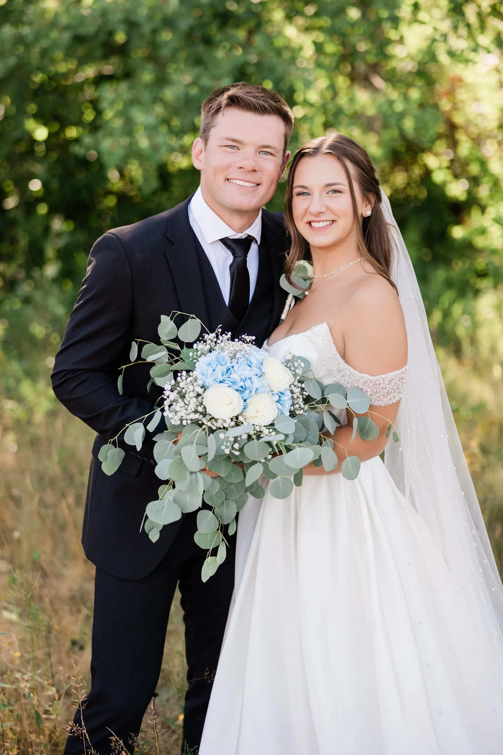 The bride and groom stand cheek to cheek smiling at the camera in a sunlit grove, her blue hydrangea and eucalyptus bouquet held between them — Tim Larsen Photography, Brainerd Lakes MN