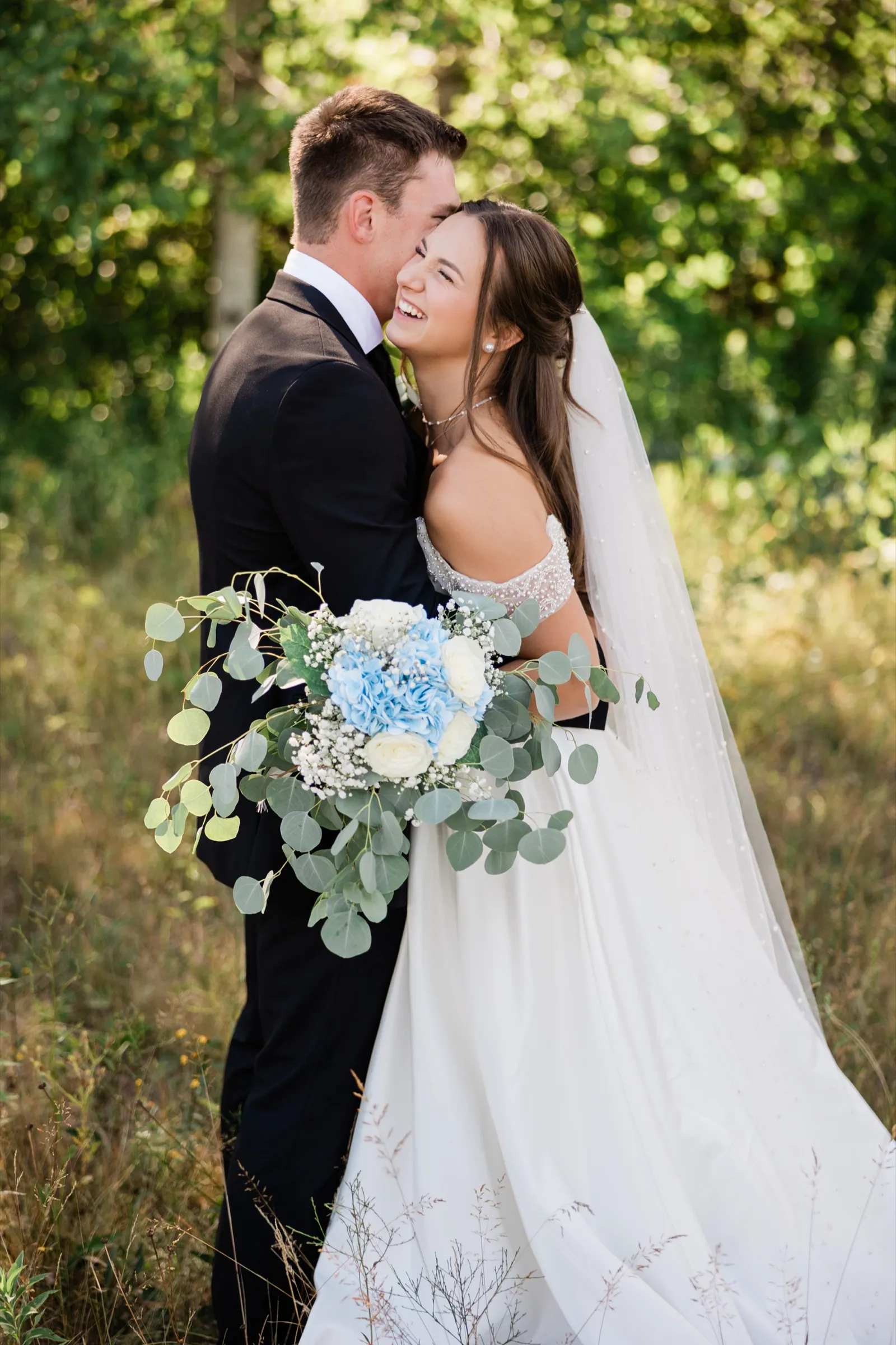The bride laughs against the groom's shoulder in knee-high grass, her veil and train spilling across the ground behind her — Tim Larsen Photography, Brainerd Lakes MN