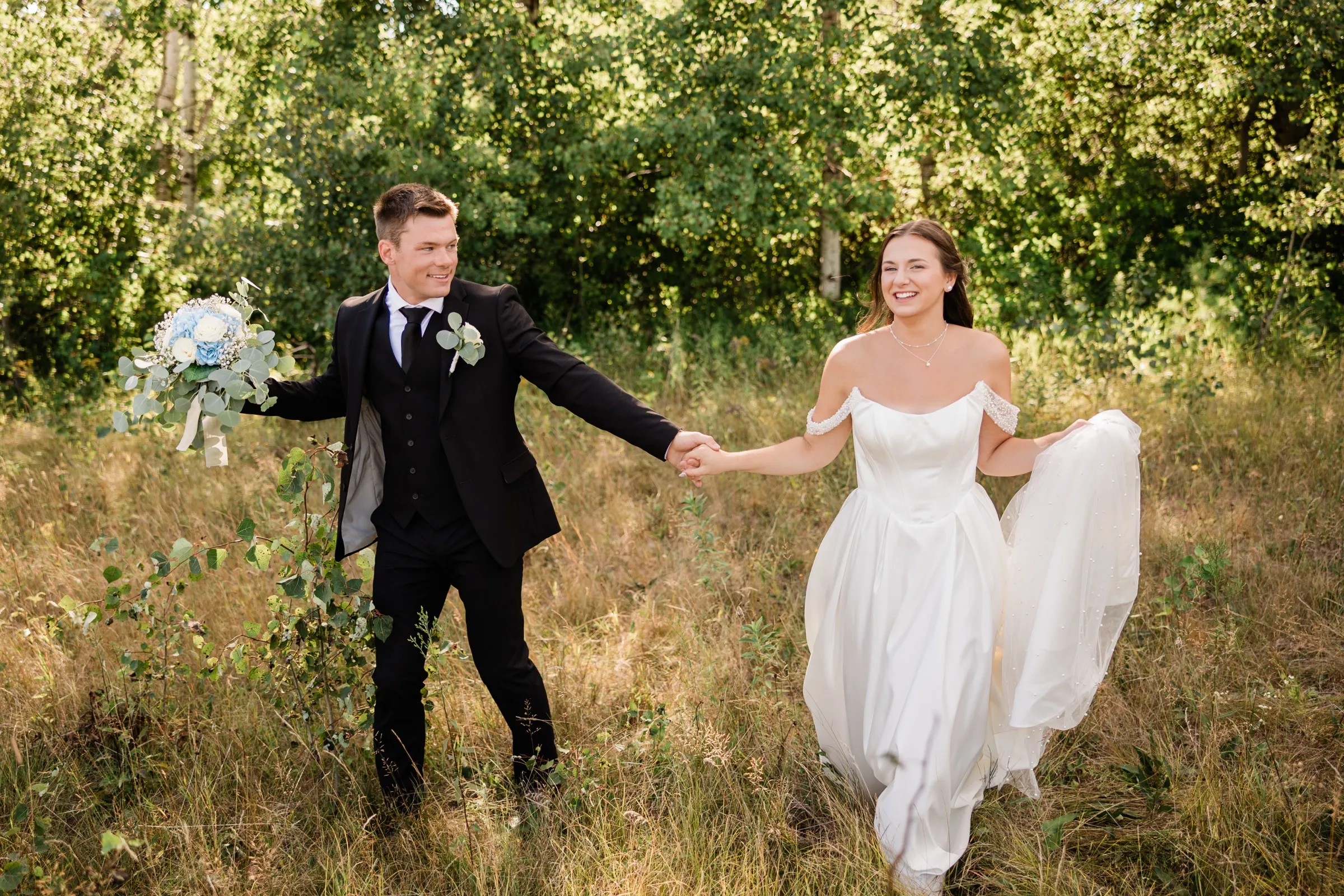 The bride and groom walk apart but hand in hand through a grassy clearing, the groom carrying her bouquet and the bride holding up her skirt — Tim Larsen Photography, Brainerd Lakes MN