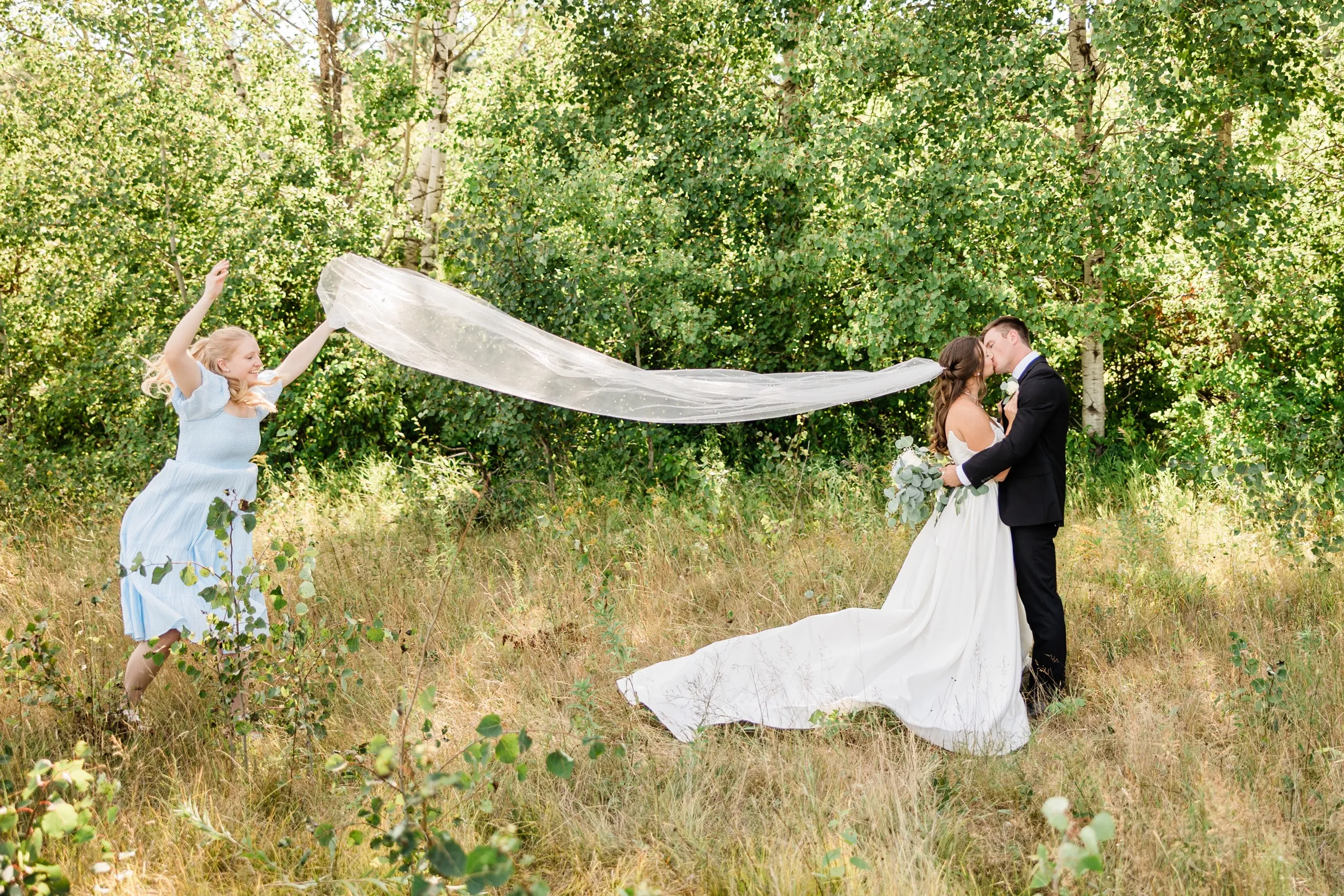 A bridesmaid in a blue dress throws the bride's long veil out sideways across a grassy clearing while the couple kisses at the other end of it — Tim Larsen Photography, Brainerd Lakes MN