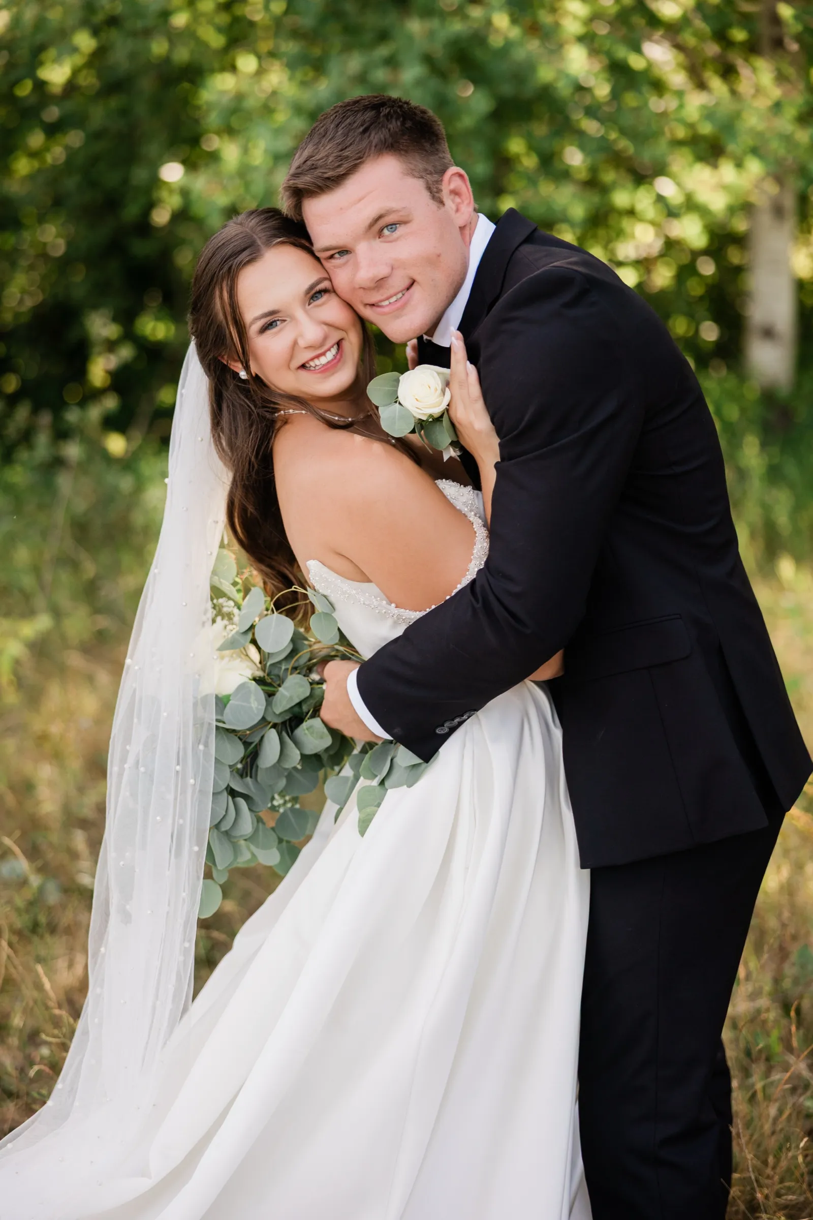 The bride and groom lean their heads together and smile at the camera, her long pearl veil falling the length of the frame to the grass — Tim Larsen Photography, Brainerd Lakes MN