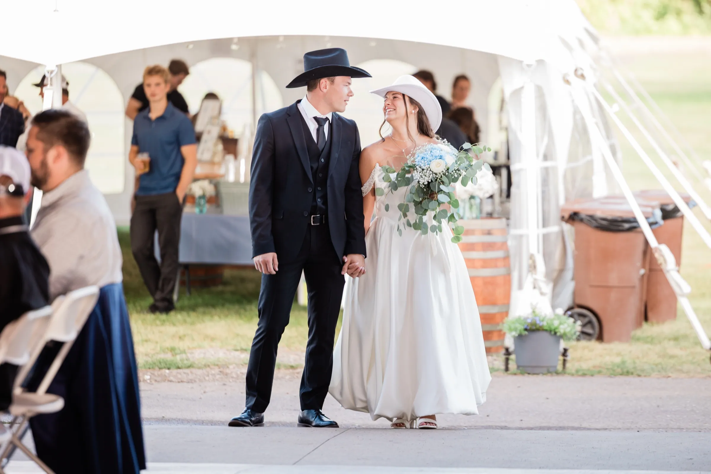 The bride in a white cowboy hat and the groom in a black one walk hand in hand past a white event tent where guests are gathered with drinks — Tim Larsen Photography, Brainerd Lakes MN