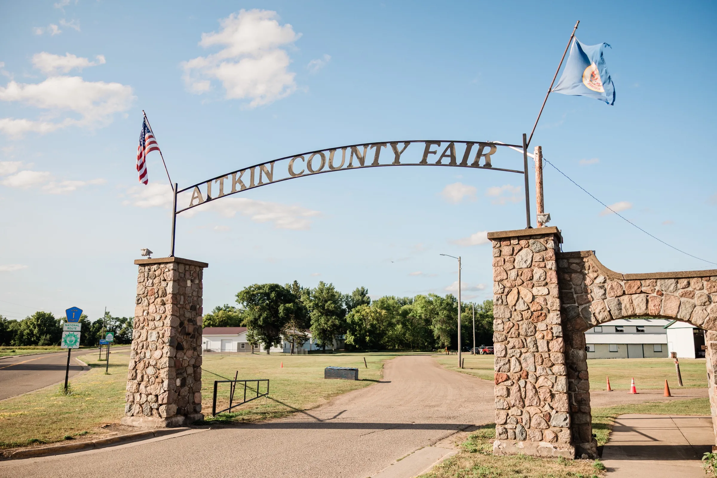 The stone entrance arch of the Aitkin County Fairgrounds reading Aitkin County Fair, with American and Minnesota flags flying — Tim Larsen Photography, Brainerd Lakes MN