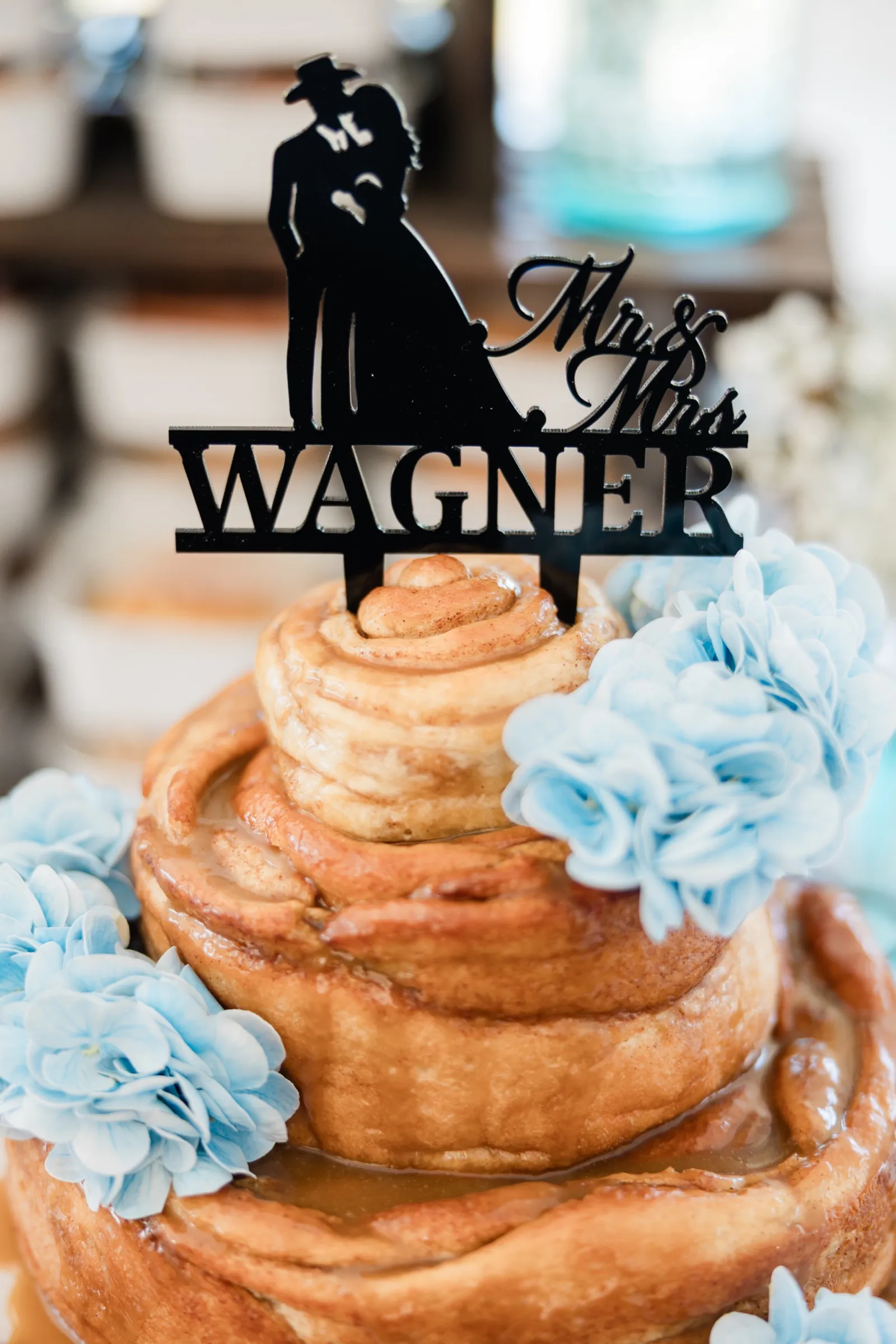 A black silhouette cake topper reading Mr. and Mrs. Wagner stands on a cinnamon roll cake trimmed with pale blue flowers — Tim Larsen Photography, Brainerd Lakes MN