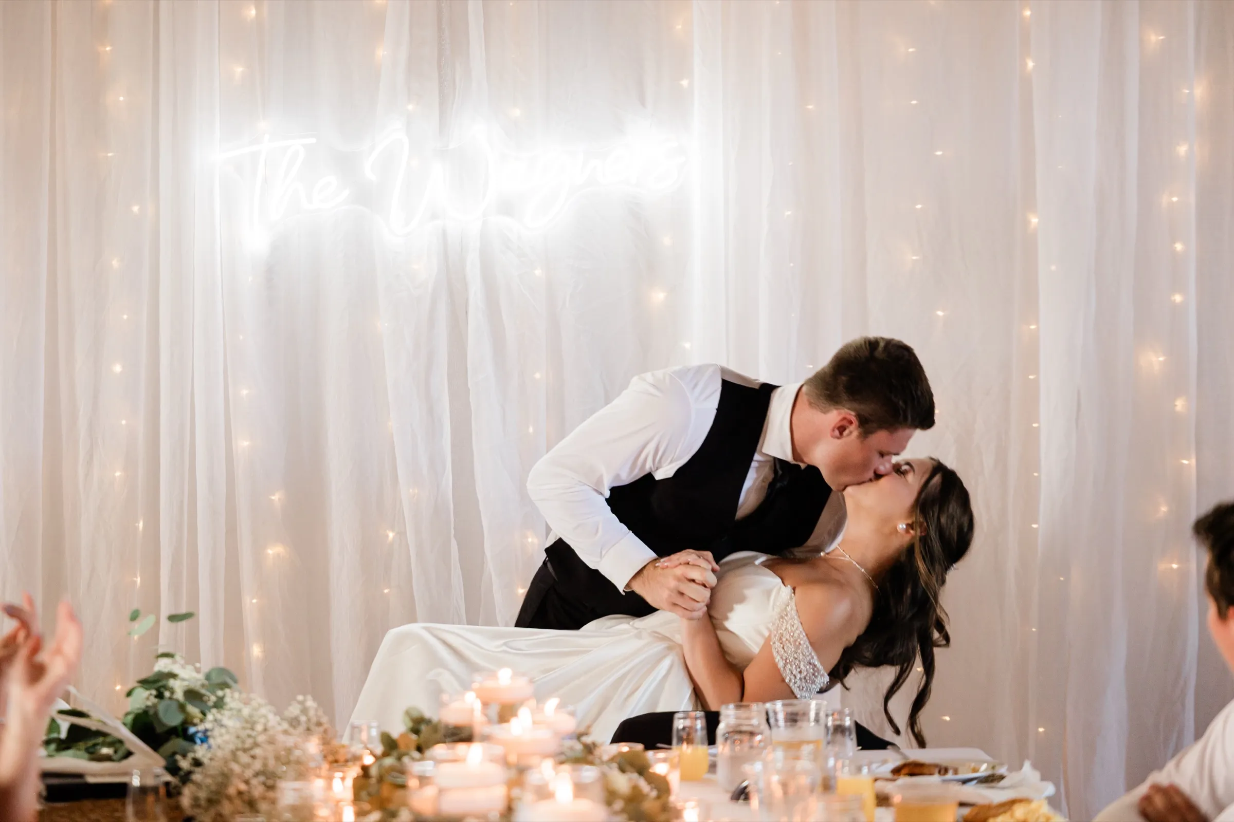 The groom dips the bride backward into a kiss at the head table beneath a glowing neon sign reading The Wagners — Tim Larsen Photography, Brainerd Lakes MN