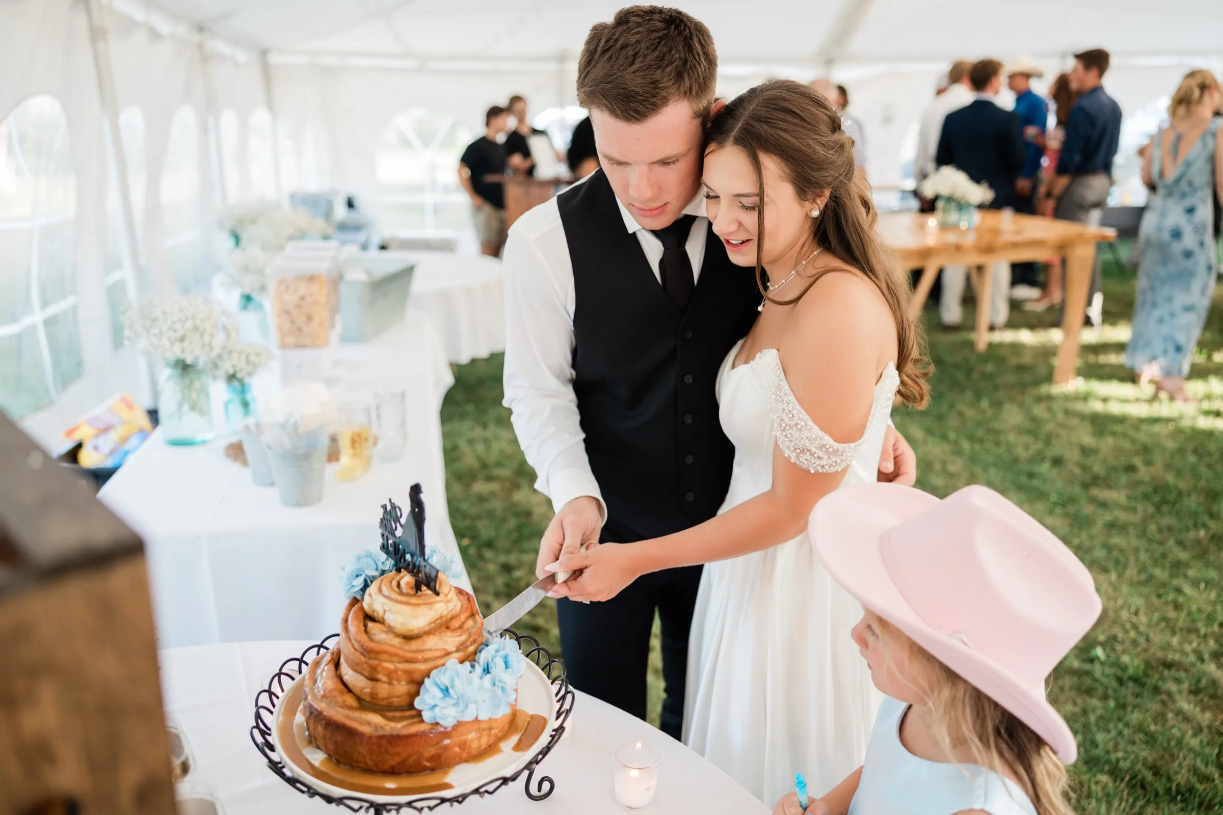 The bride and groom lean their heads together as they cut into a cinnamon roll cake while a girl in a pink cowboy hat watches — Tim Larsen Photography, Brainerd Lakes MN