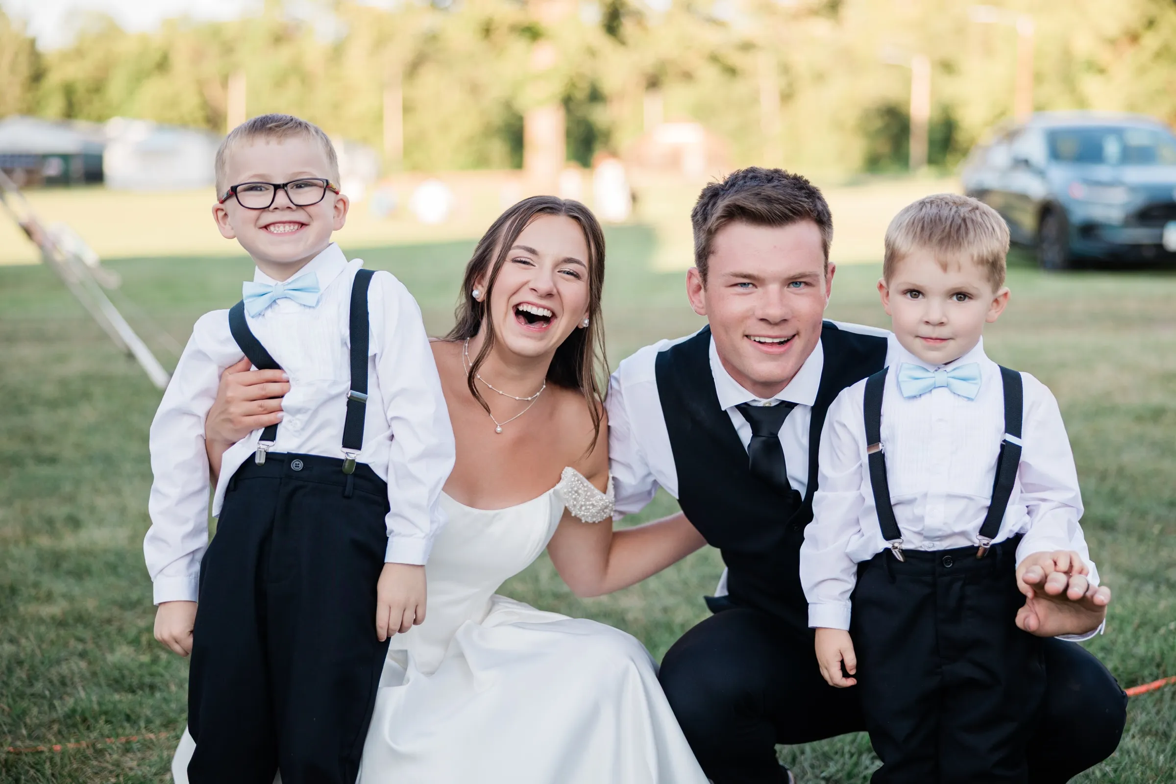 The bride and groom crouch on the grass between two young ring bearers in suspenders and bow ties, all four laughing — Tim Larsen Photography, Brainerd Lakes MN