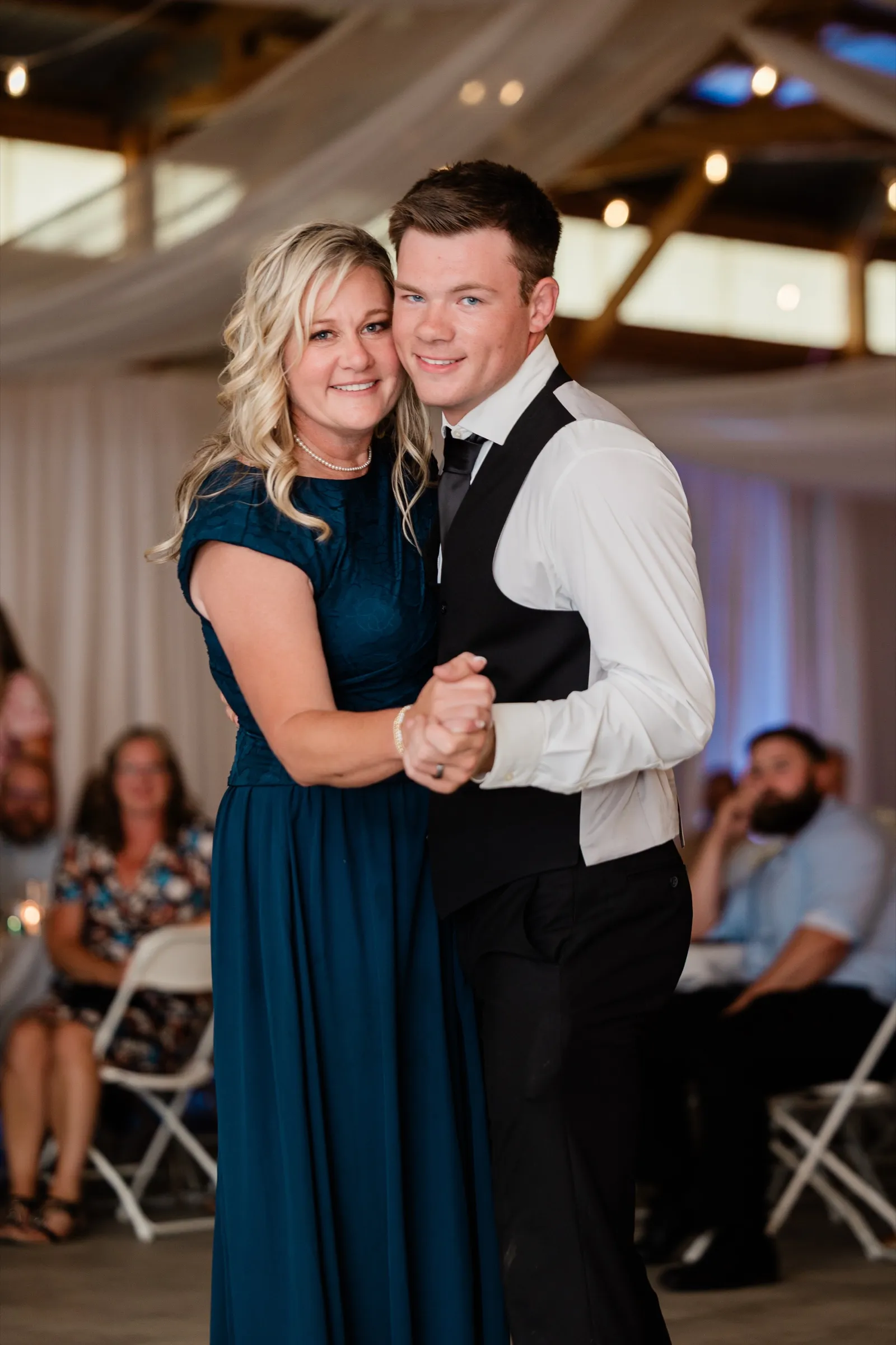 The groom dances with his mother in a teal gown, both smiling toward the camera under string lights in the reception hall — Tim Larsen Photography, Brainerd Lakes MN