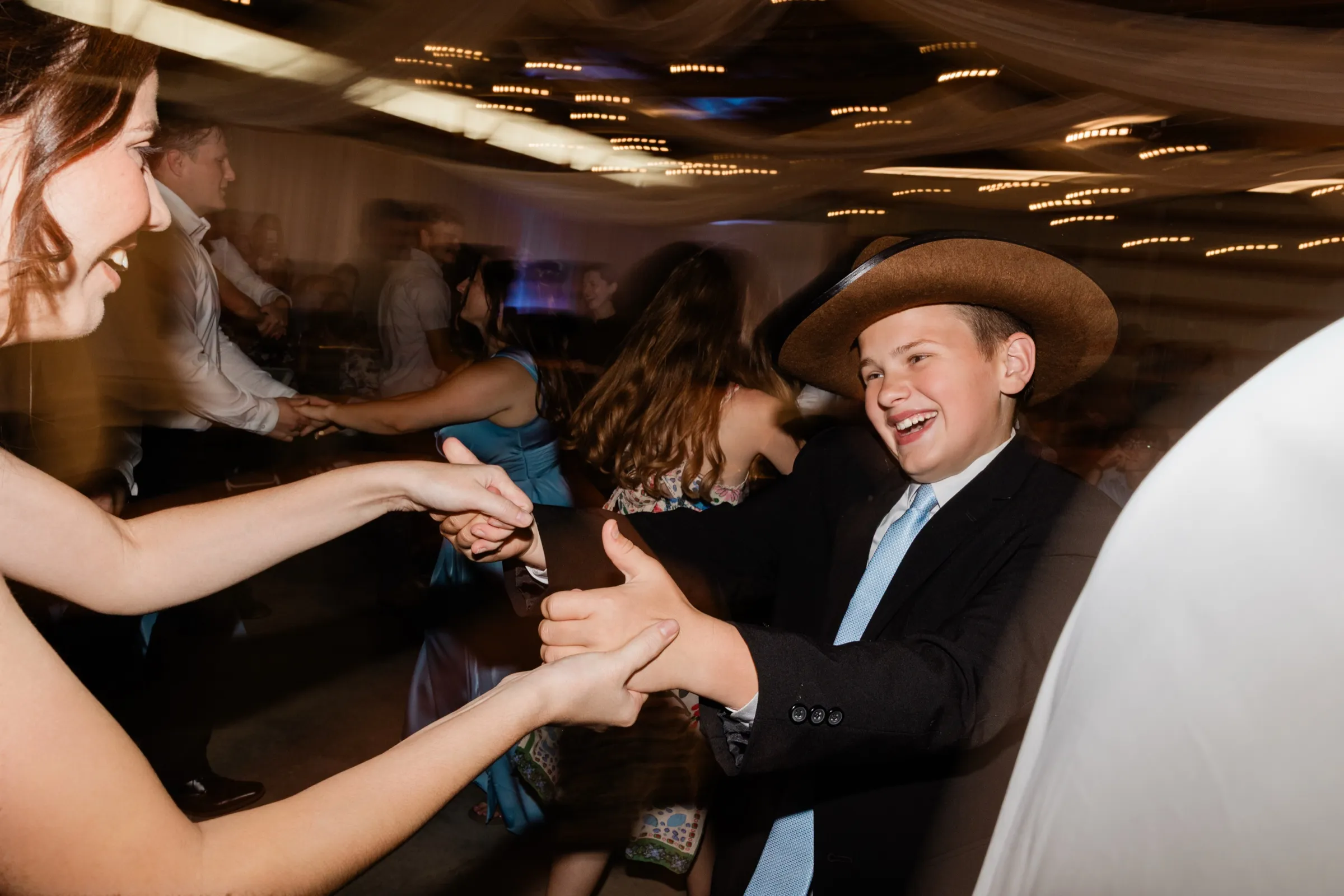 A boy in a brown cowboy hat and dark suit laughs while joining hands with a woman at the edge of the frame, the rest of the dance floor blurred by motion — Tim Larsen Photography, Brainerd Lakes MN