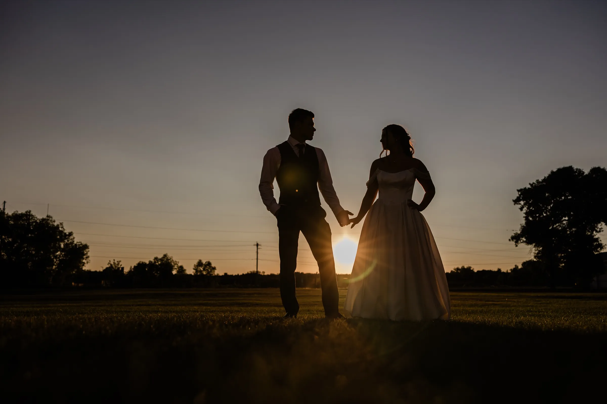 The bride and groom stand in silhouette facing each other on the open Aitkin County Fairgrounds field at sunset — Tim Larsen Photography, Brainerd Lakes MN