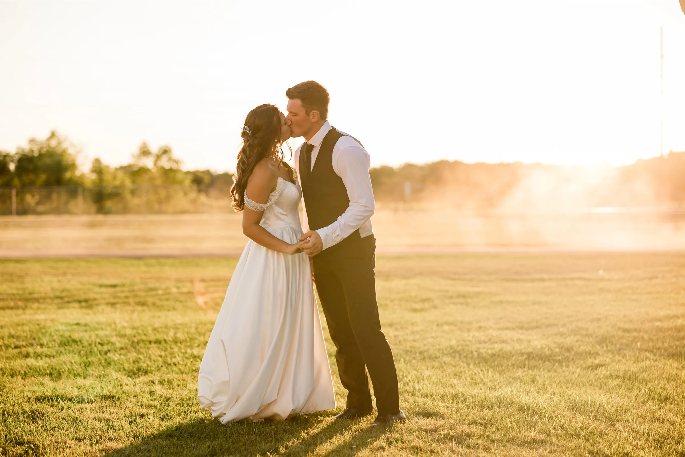 The bride and groom kiss while standing in an open field of mown grass, their joined hands between them, as low backlight washes the right side of the frame into haze — Tim Larsen Photography, Brainerd Lakes MN