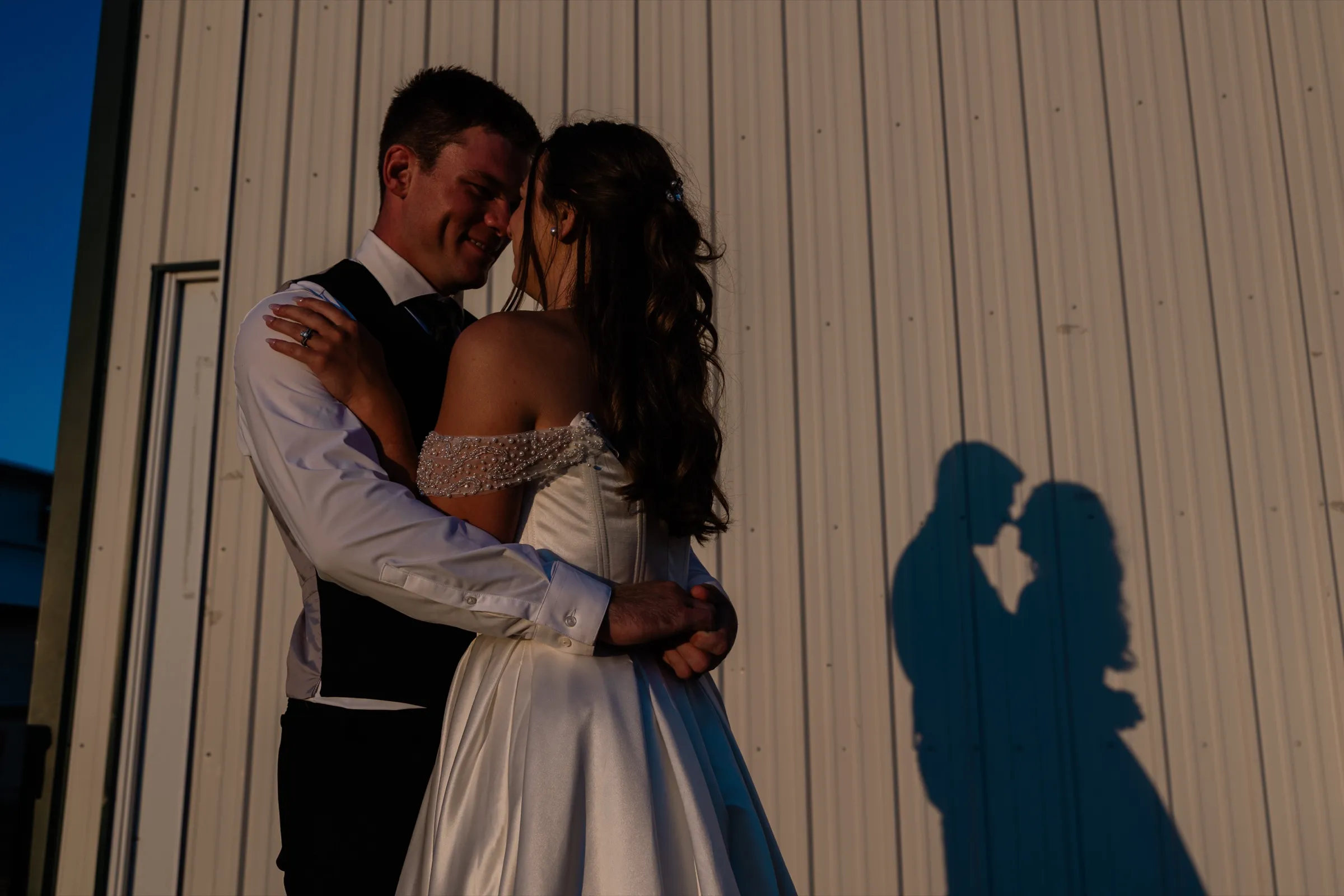 The bride and groom lean in for a kiss against a white fair building at the Aitkin County Fairgrounds, their embrace cast as a shadow beside them — Tim Larsen Photography, Brainerd Lakes MN