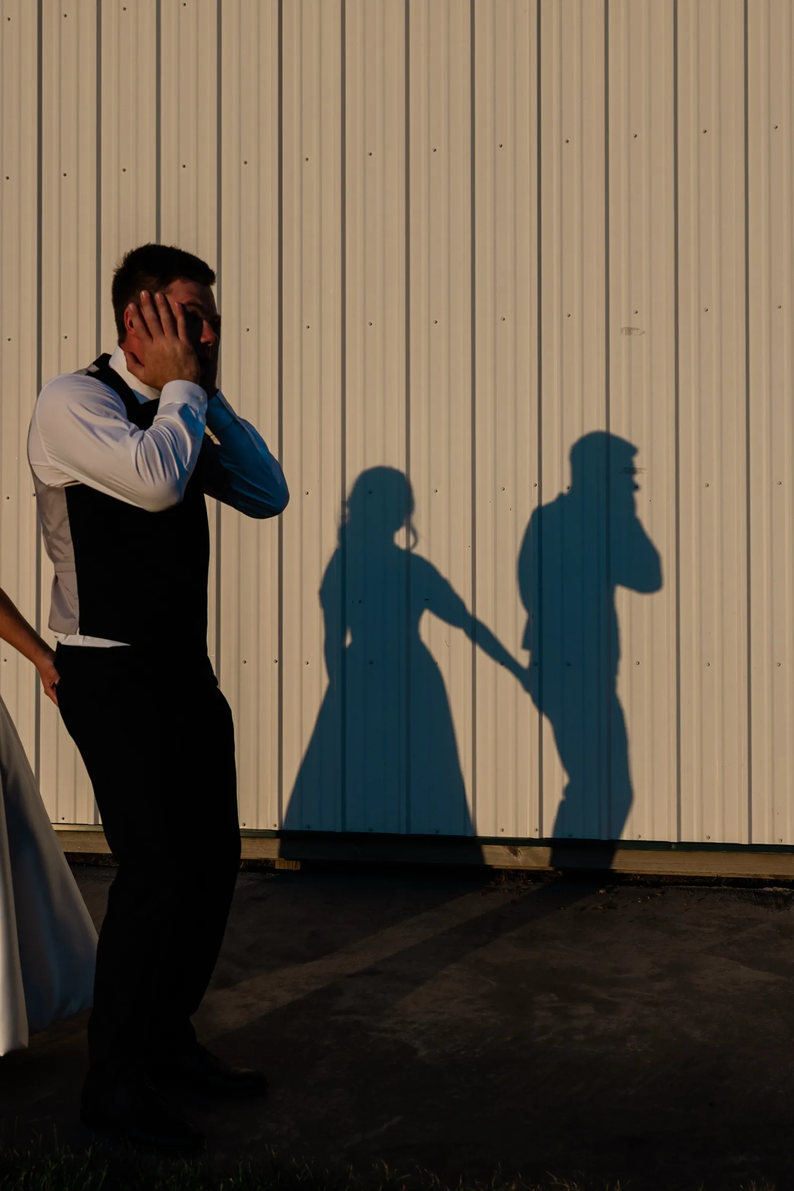 The groom stands against a white corrugated metal building with both hands over his eyes while the shadows of the couple holding hands stretch across the siding beside him — Tim Larsen Photography, Brainerd Lakes MN