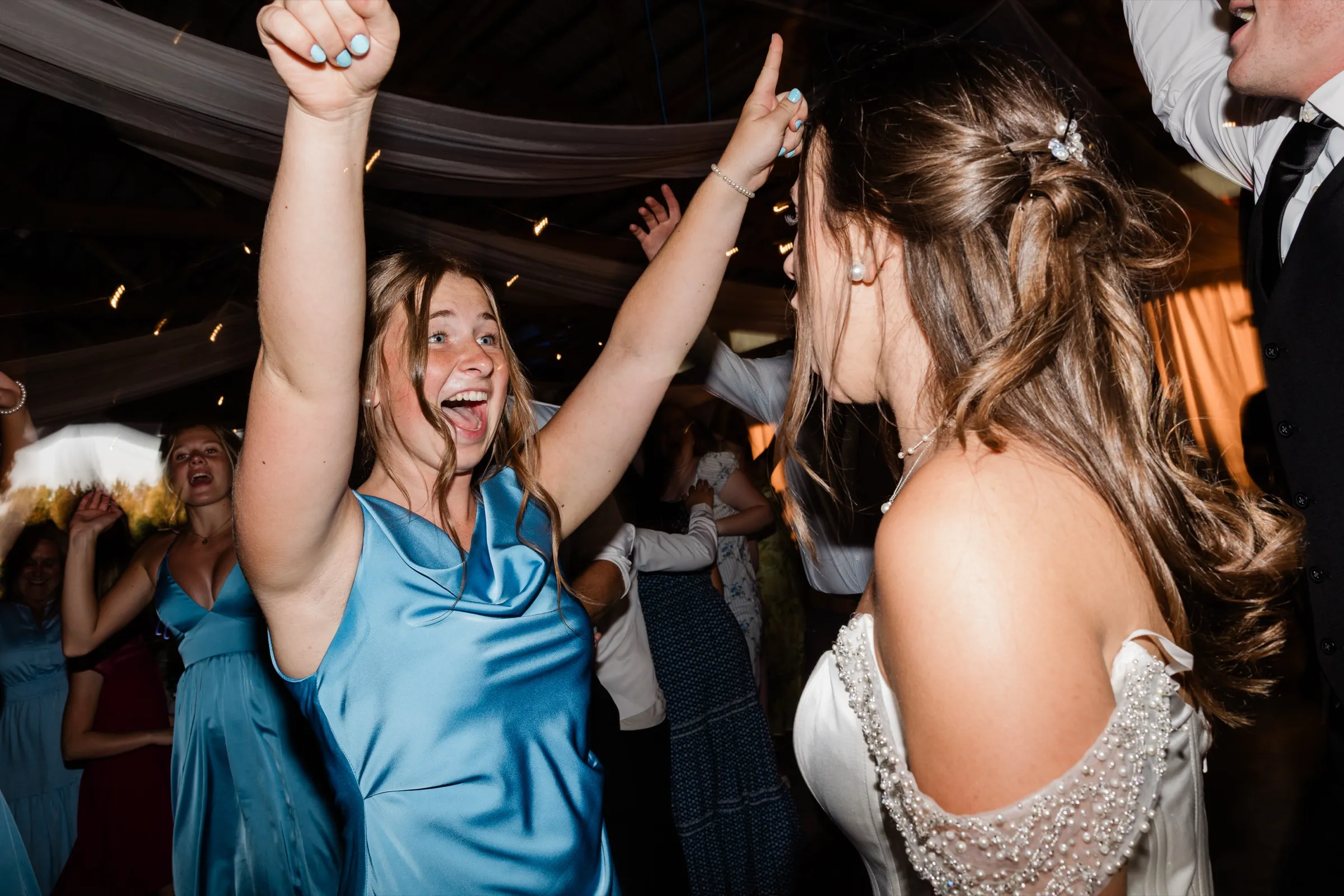 A bridesmaid in a blue satin dress throws both arms overhead and shouts the words of a song directly at the bride, who is seen from behind in her pearl-trimmed gown — Tim Larsen Photography, Brainerd Lakes MN