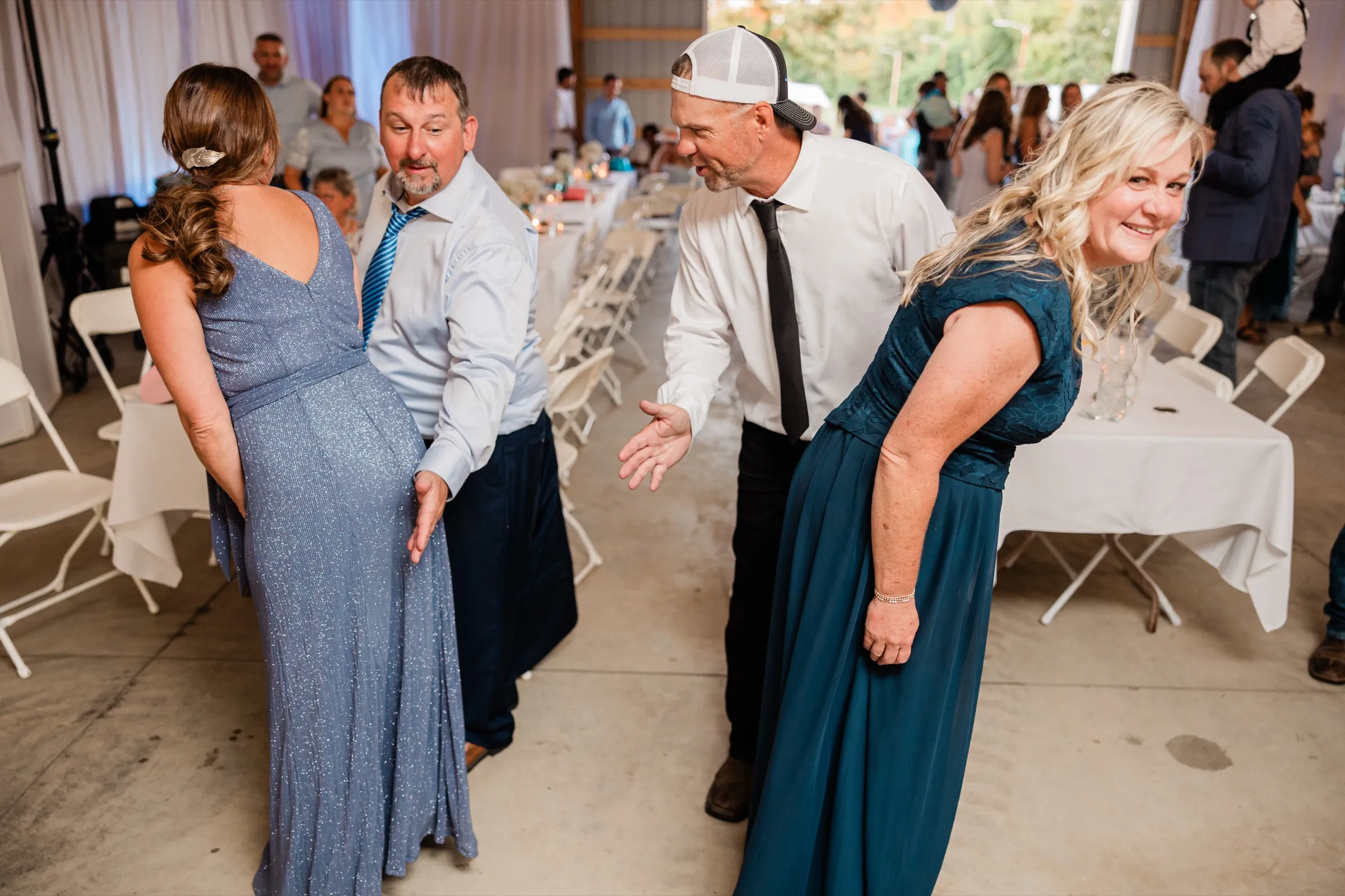 Two couples bend forward into an exaggerated dance move on the concrete floor beside long white banquet tables, one woman grinning back over her shoulder at the camera — Tim Larsen Photography, Brainerd Lakes MN