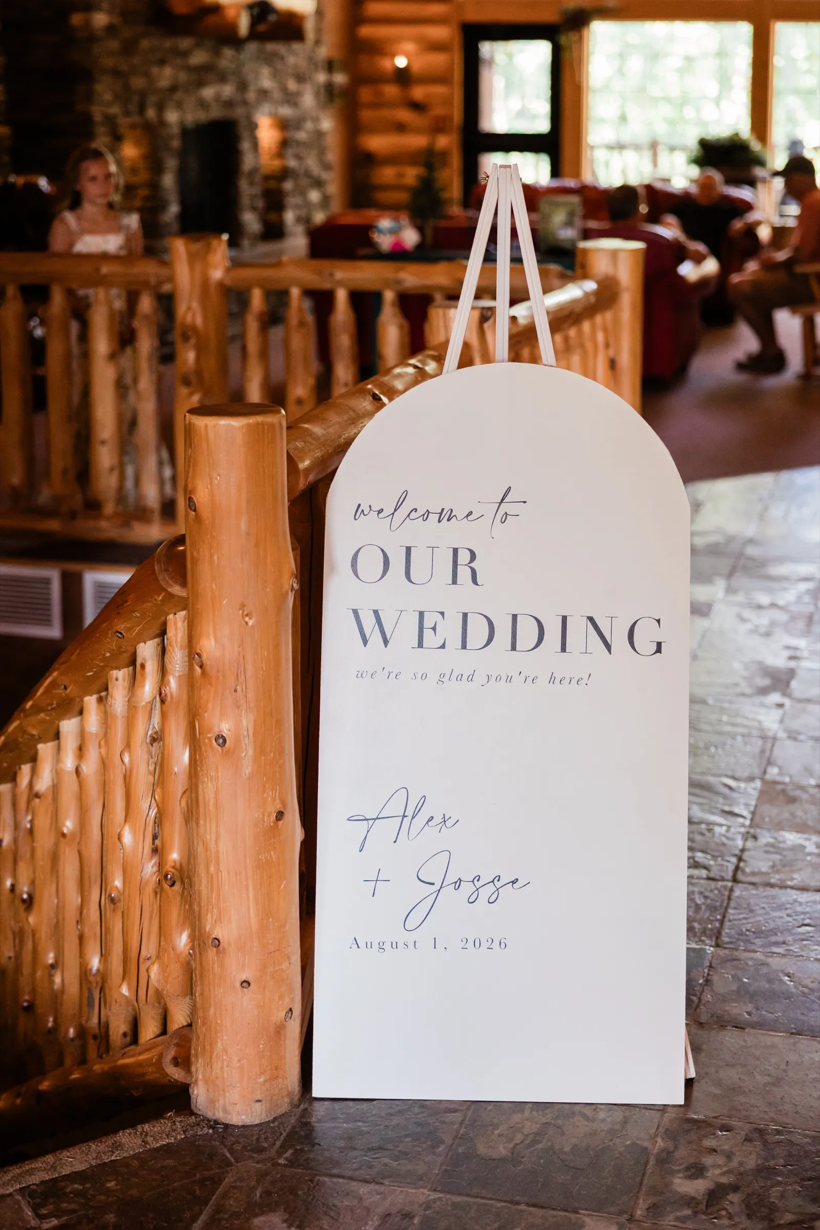An arched white welcome sign reading 'Welcome to Our Wedding, Alex + Josse, August 1, 2026' stands on an easel beside a peeled-log staircase railing in the Whitefish Lodge lobby — Tim Larsen Photography, Brainerd Lakes MN