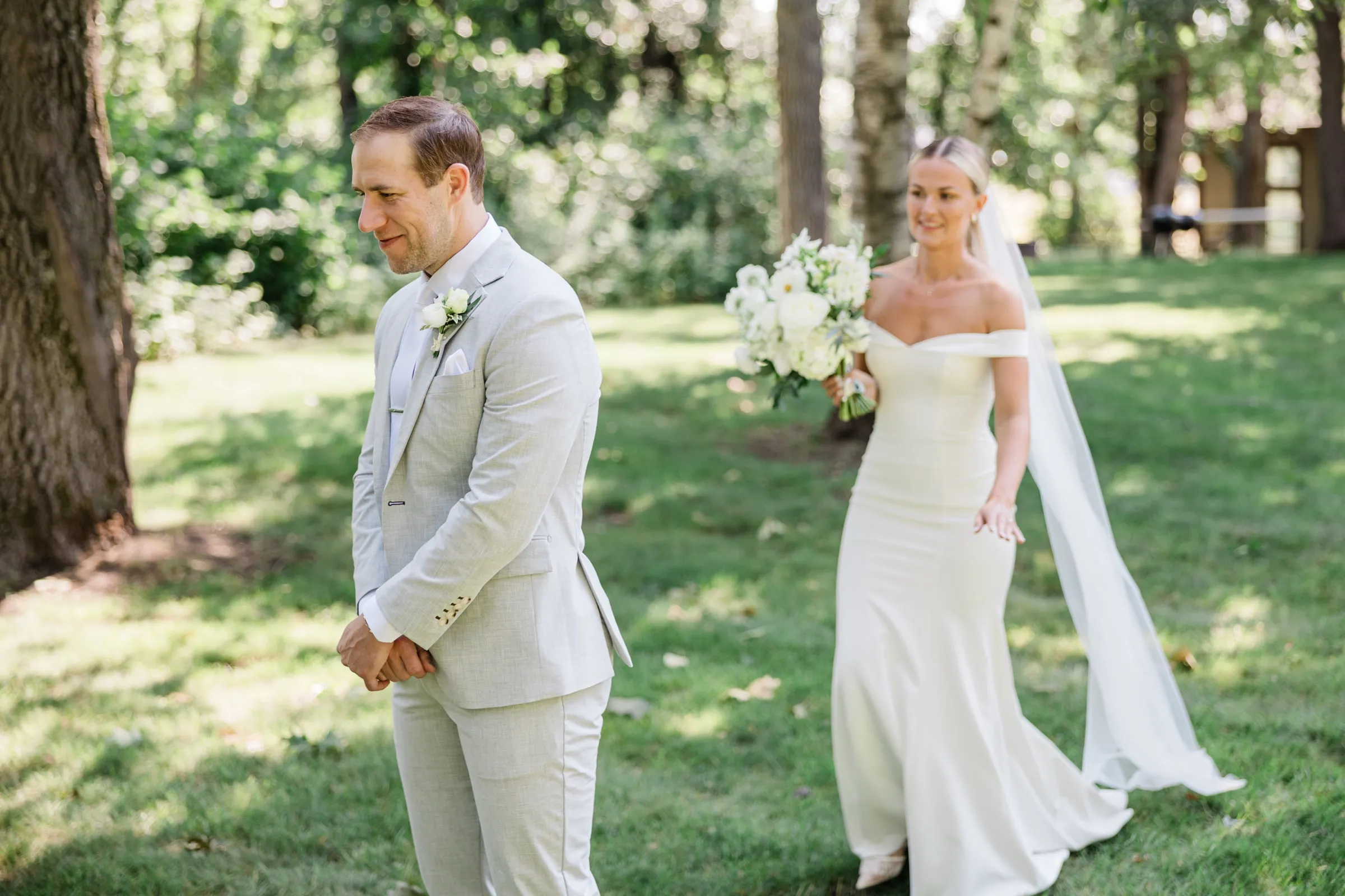 Alex stands with hands clasped and eyes lowered as Josse walks toward him across a shaded lawn for their first look — Tim Larsen Photography, Brainerd Lakes MN