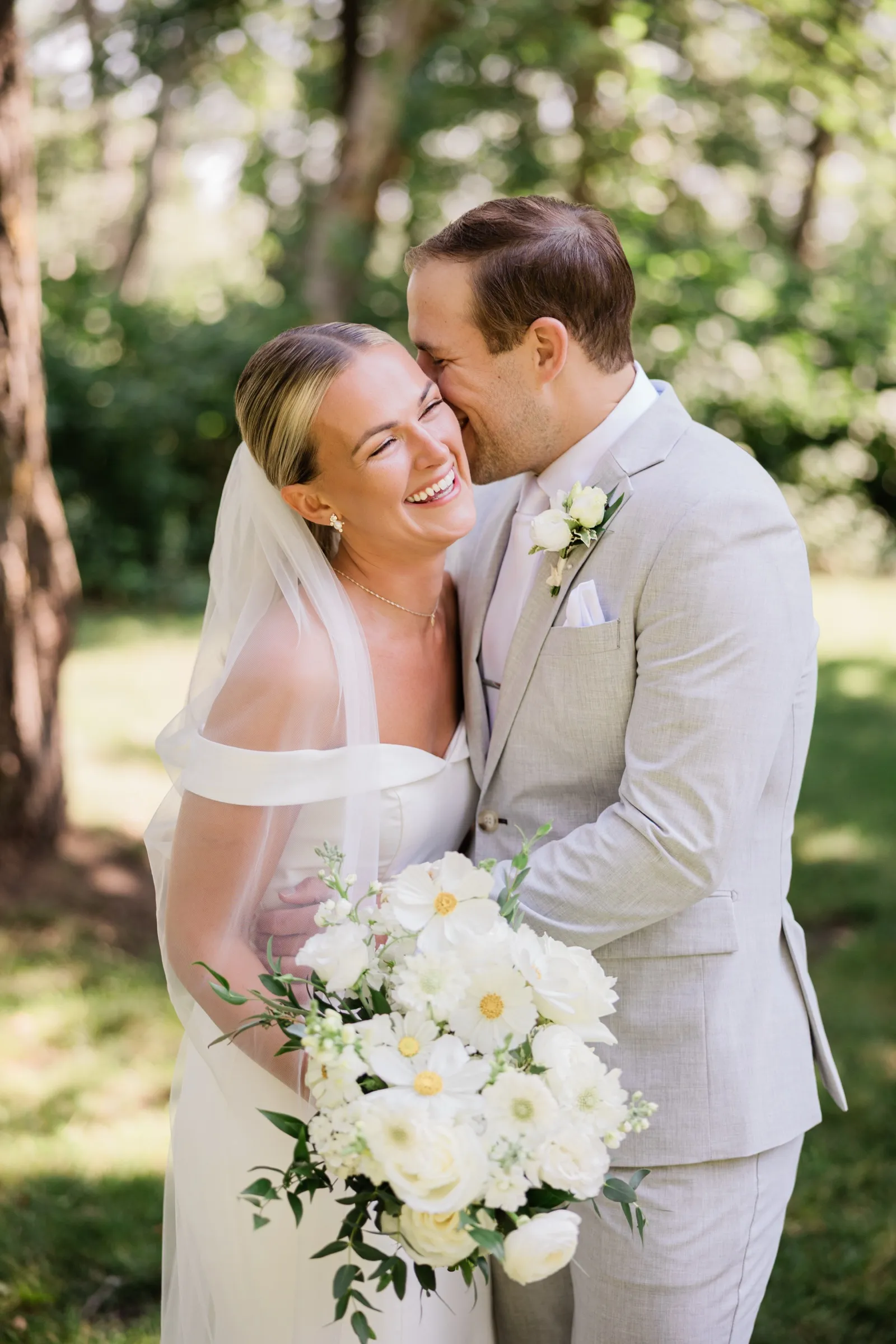 Josse laughs with her eyes closed as Alex leans in to kiss her cheek on a shaded lawn — Tim Larsen Photography, Brainerd Lakes MN