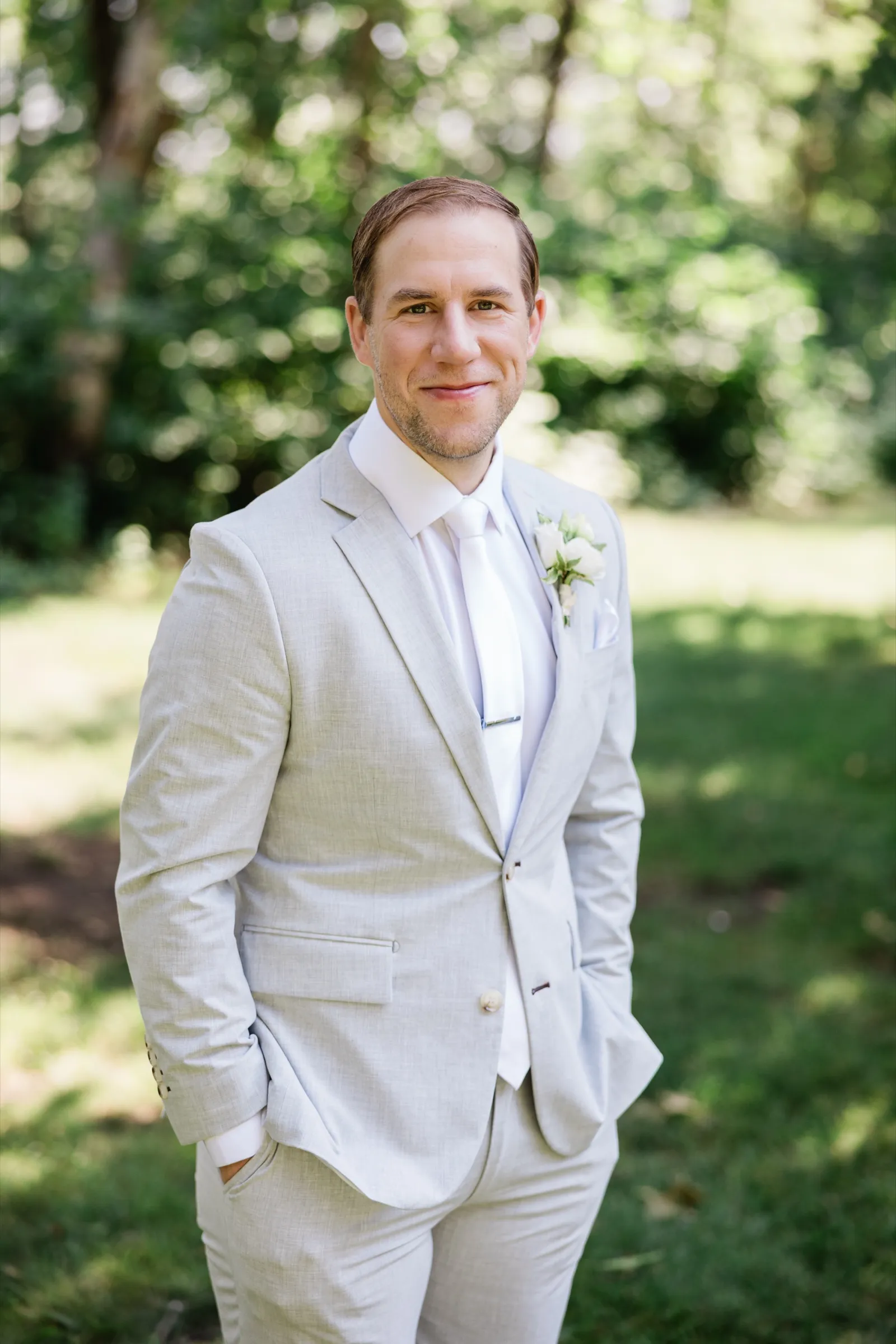 Alex stands with hands in his pockets in a light gray suit and white tie on the shaded Whitefish Lodge grounds — Tim Larsen Photography, Brainerd Lakes MN