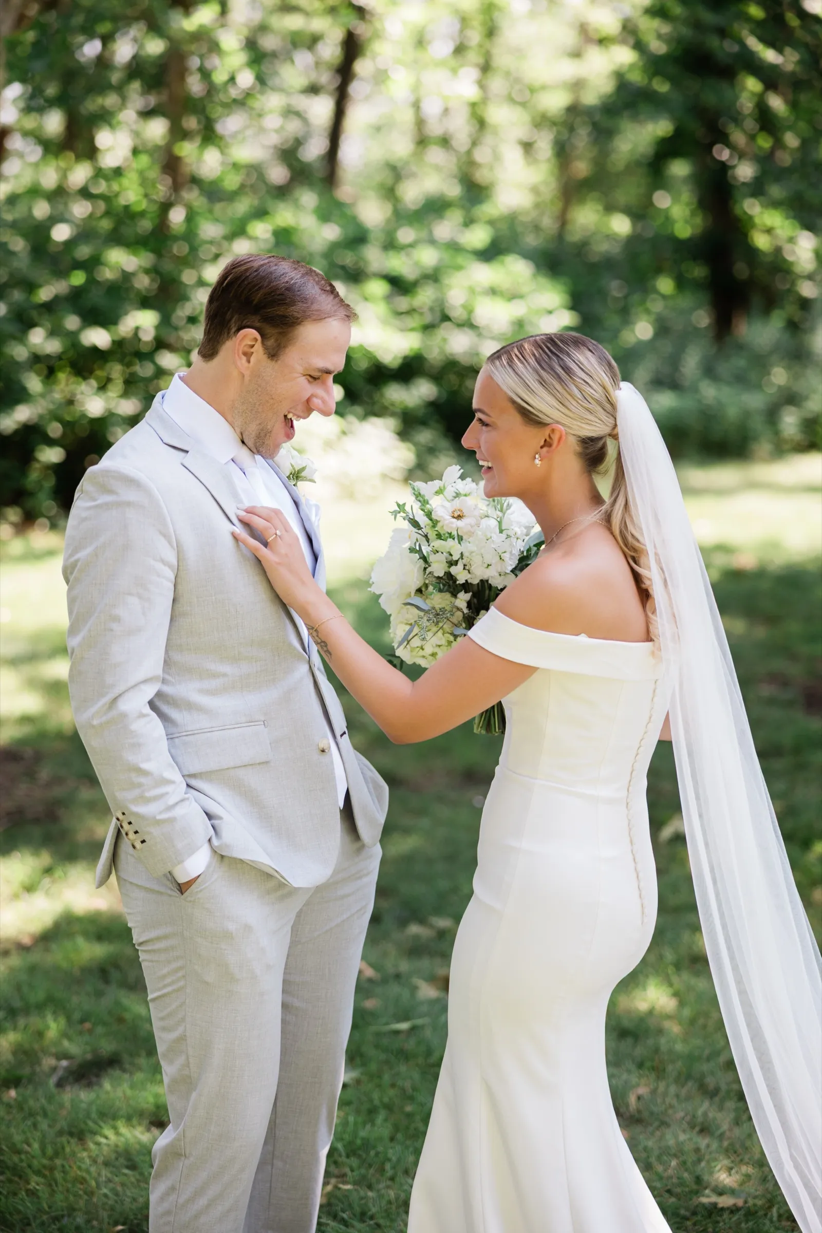 Josse rests a hand on Alex's chest as both laugh facing each other on a shaded green lawn, her veil trailing behind her — Tim Larsen Photography, Brainerd Lakes MN