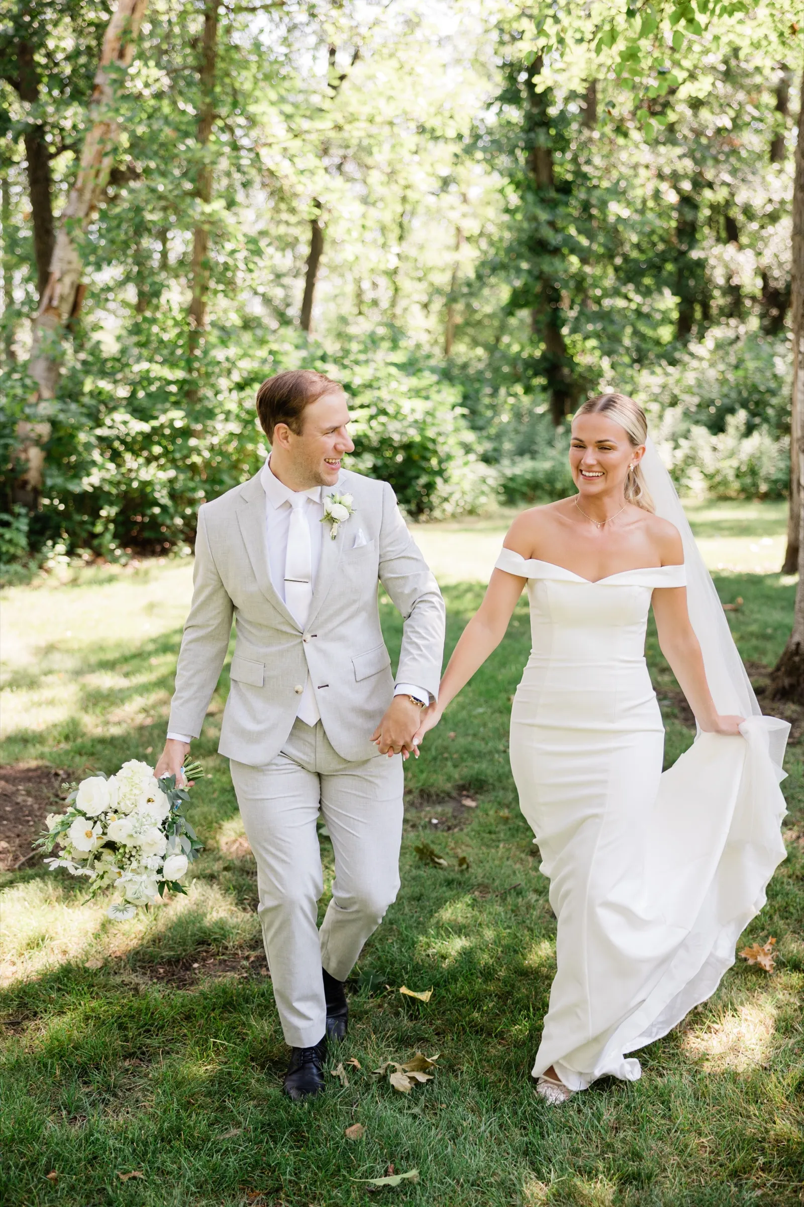 Josse holds the train of her gown as she and Alex walk hand in hand across a shaded lawn, both laughing — Tim Larsen Photography, Brainerd Lakes MN