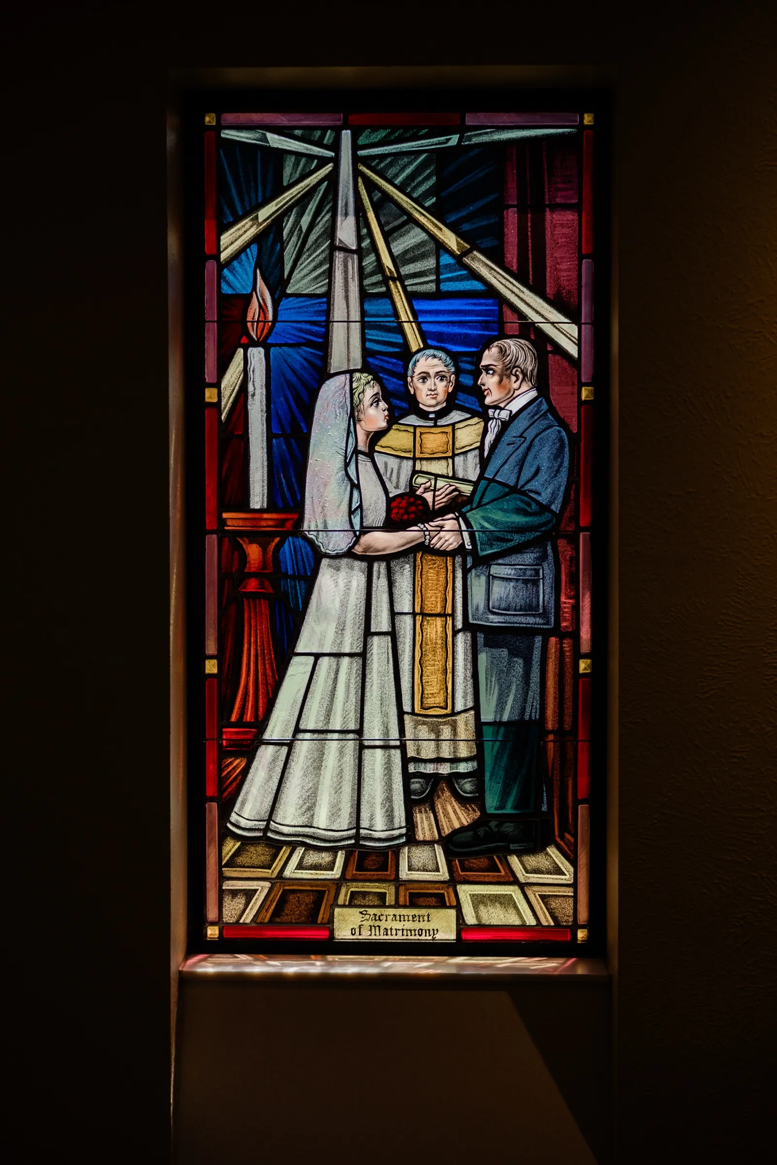 A backlit stained glass window titled 'Sacrament of Matrimony' depicting a bride and groom joining hands before a priest — Tim Larsen Photography, Brainerd Lakes MN