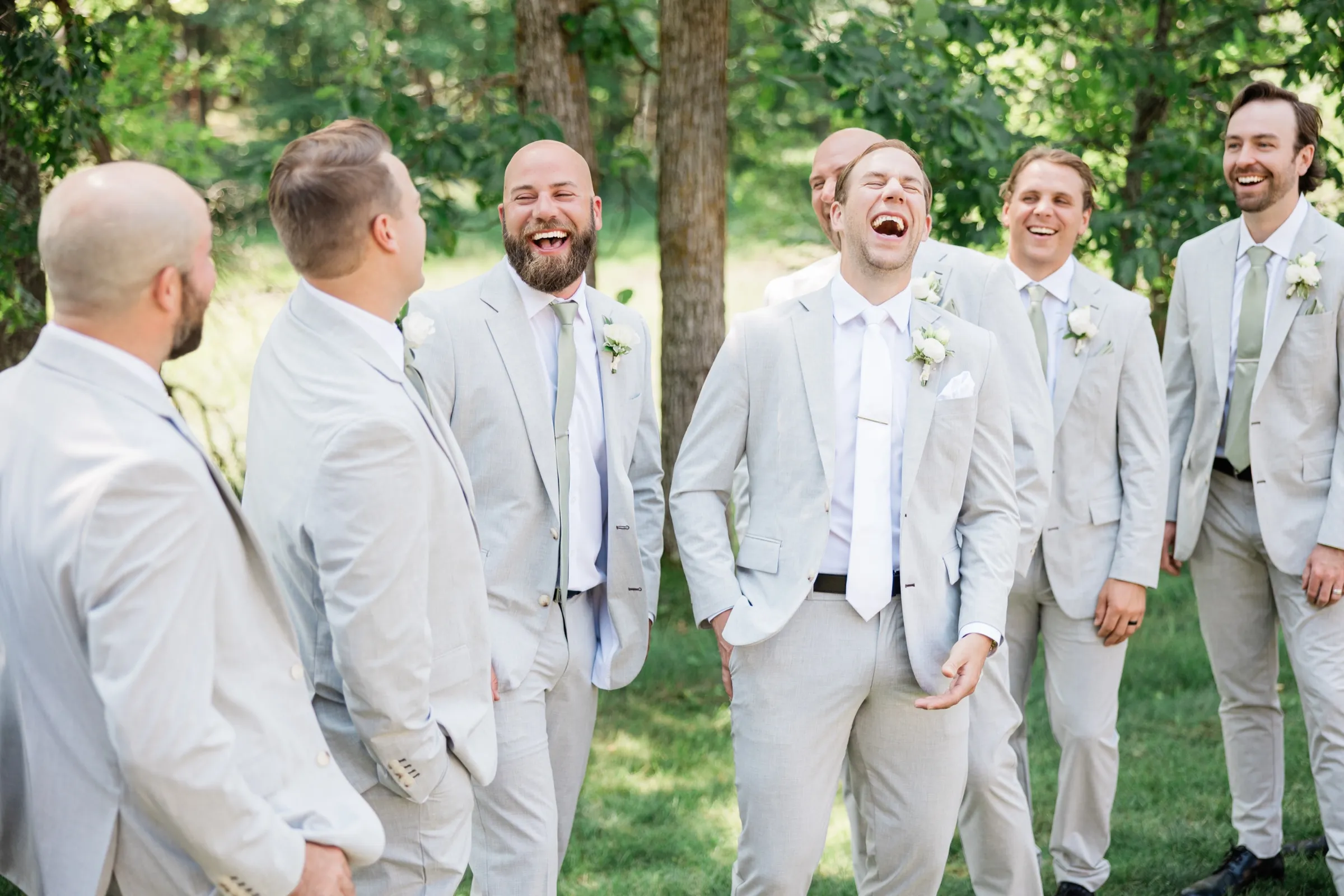 Alex throws his head back laughing with six groomsmen in light gray suits and sage ties on a shaded lawn — Tim Larsen Photography, Brainerd Lakes MN