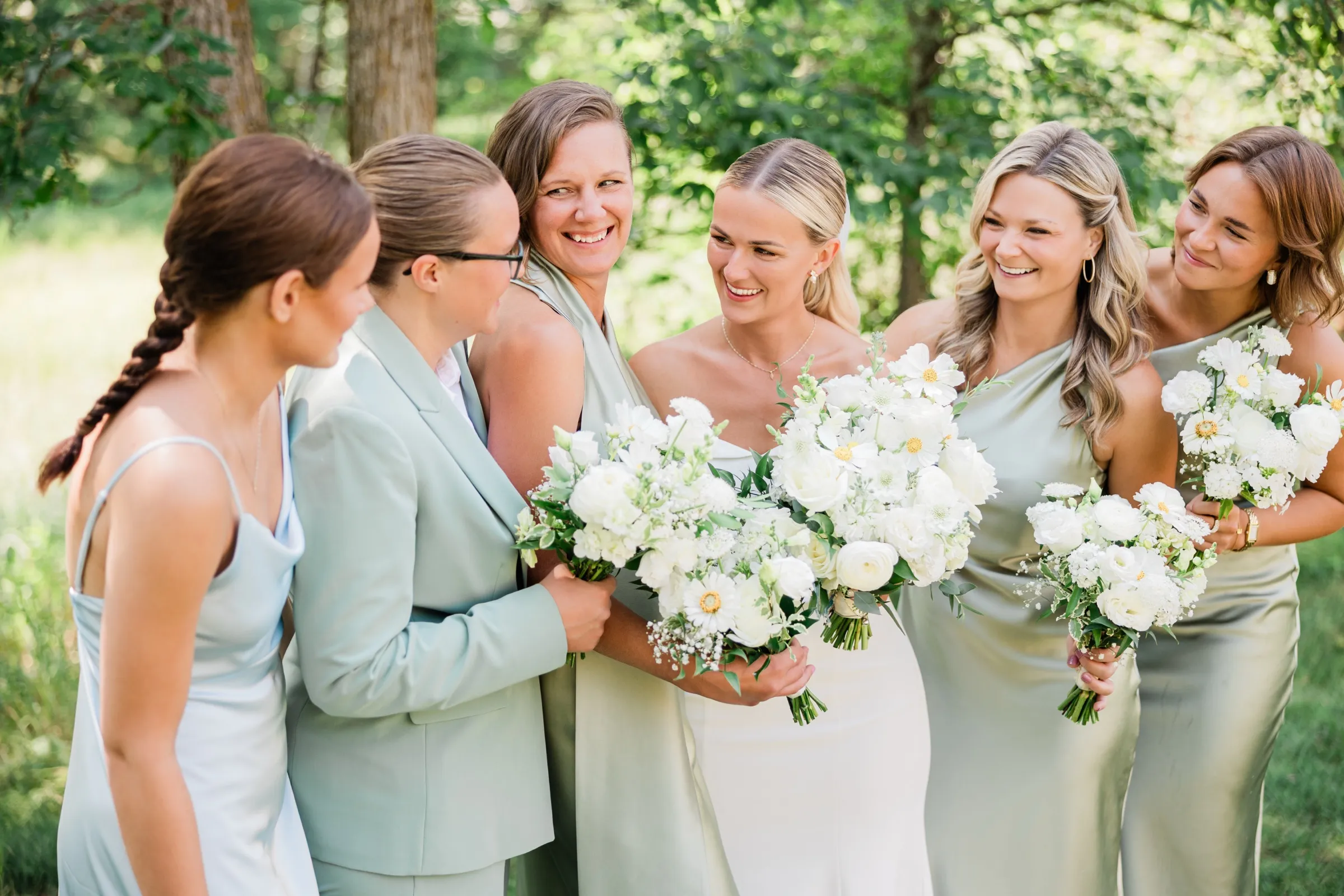 Josse laughs with her bridesmaids in sage green dresses as they gather close holding white bouquets in a wooded clearing — Tim Larsen Photography, Brainerd Lakes MN