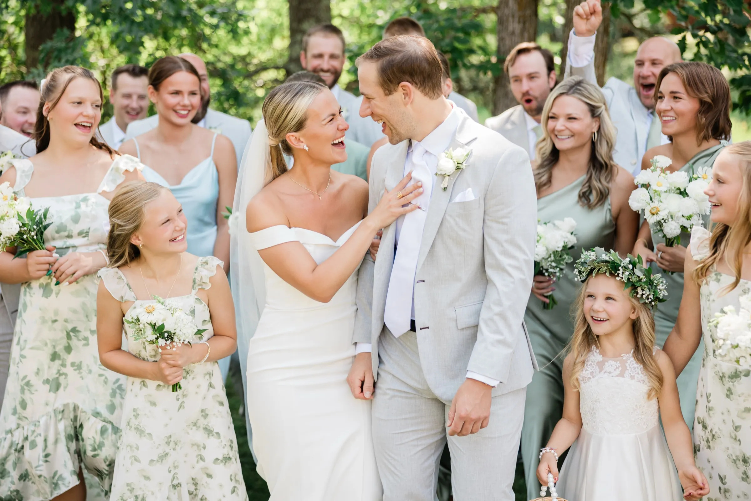 Josse rests a hand on Alex's chest as they laugh face to face, surrounded by a cheering bridal party and children in sage and floral dresses — Tim Larsen Photography, Brainerd Lakes MN