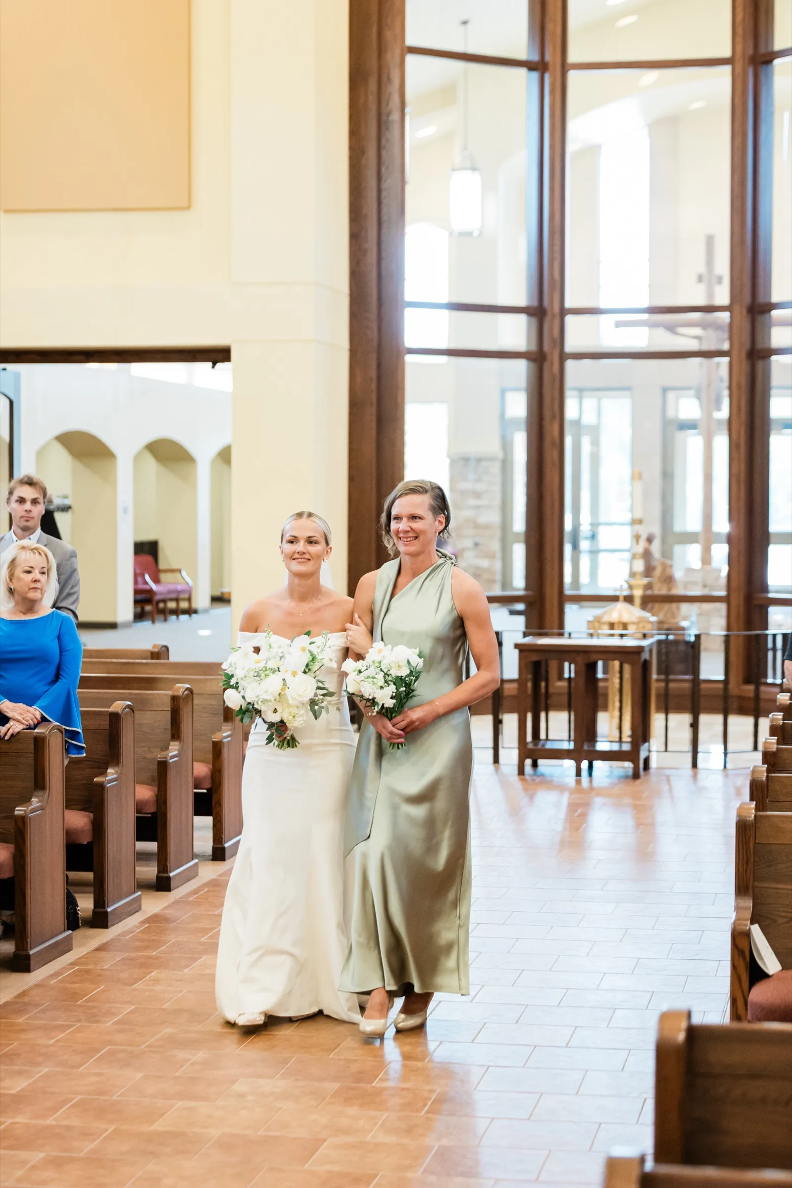 Josse walks arm in arm with her mother down the aisle of Immaculate Heart Catholic Church in Crosslake, both holding white bouquets — Tim Larsen Photography, Brainerd Lakes MN