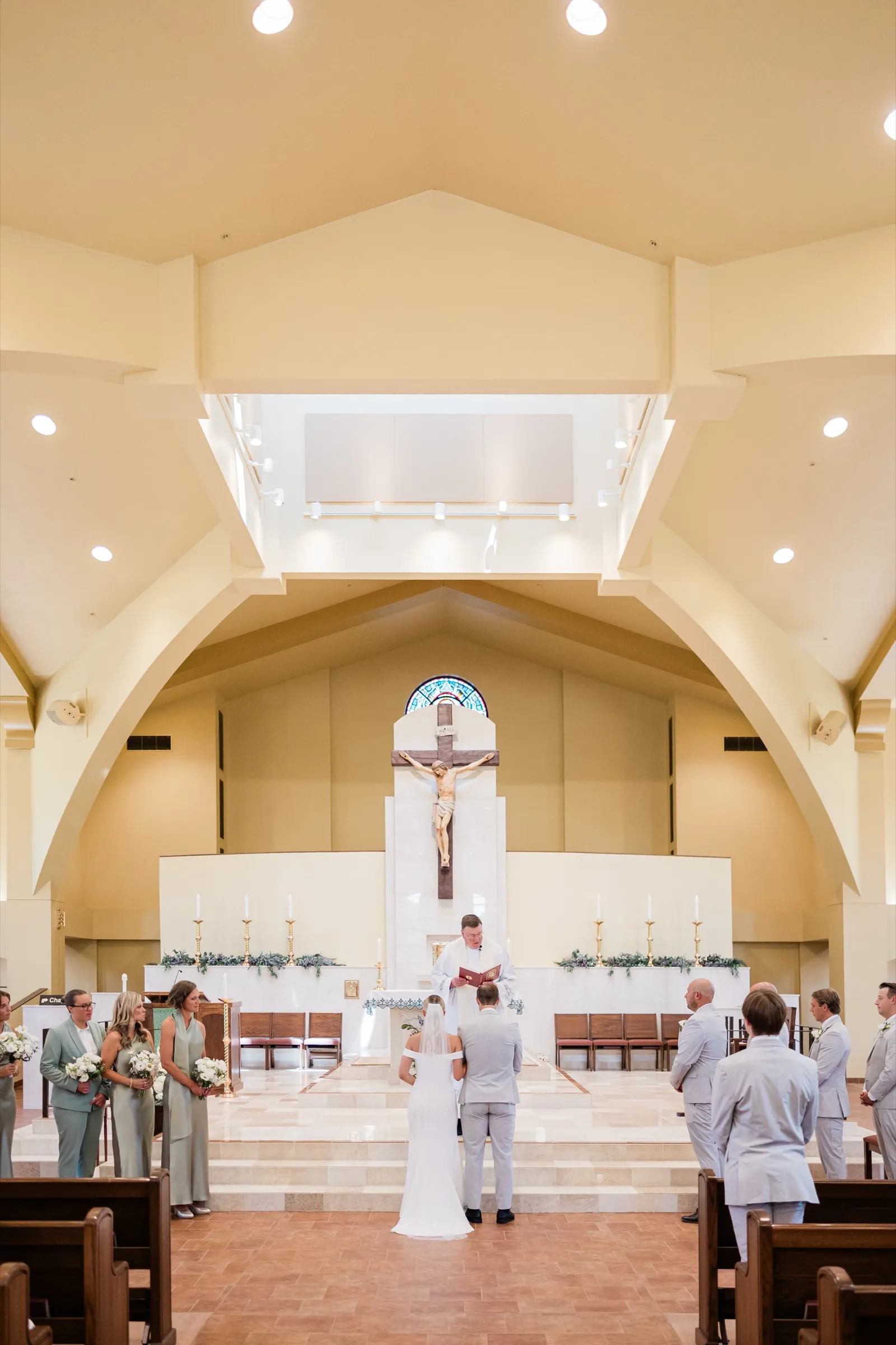 Josse and Alex stand facing the priest at the altar steps of the light-filled church, flanked by their bridal party beneath a large crucifix — Tim Larsen Photography, Brainerd Lakes MN