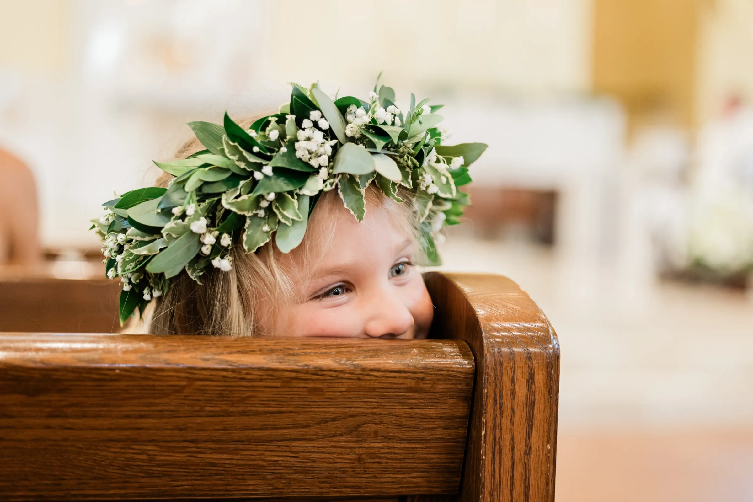 A flower girl in a greenery and baby's breath crown peeks over the back of a wooden church pew, one eye above the rail — Tim Larsen Photography, Brainerd Lakes MN