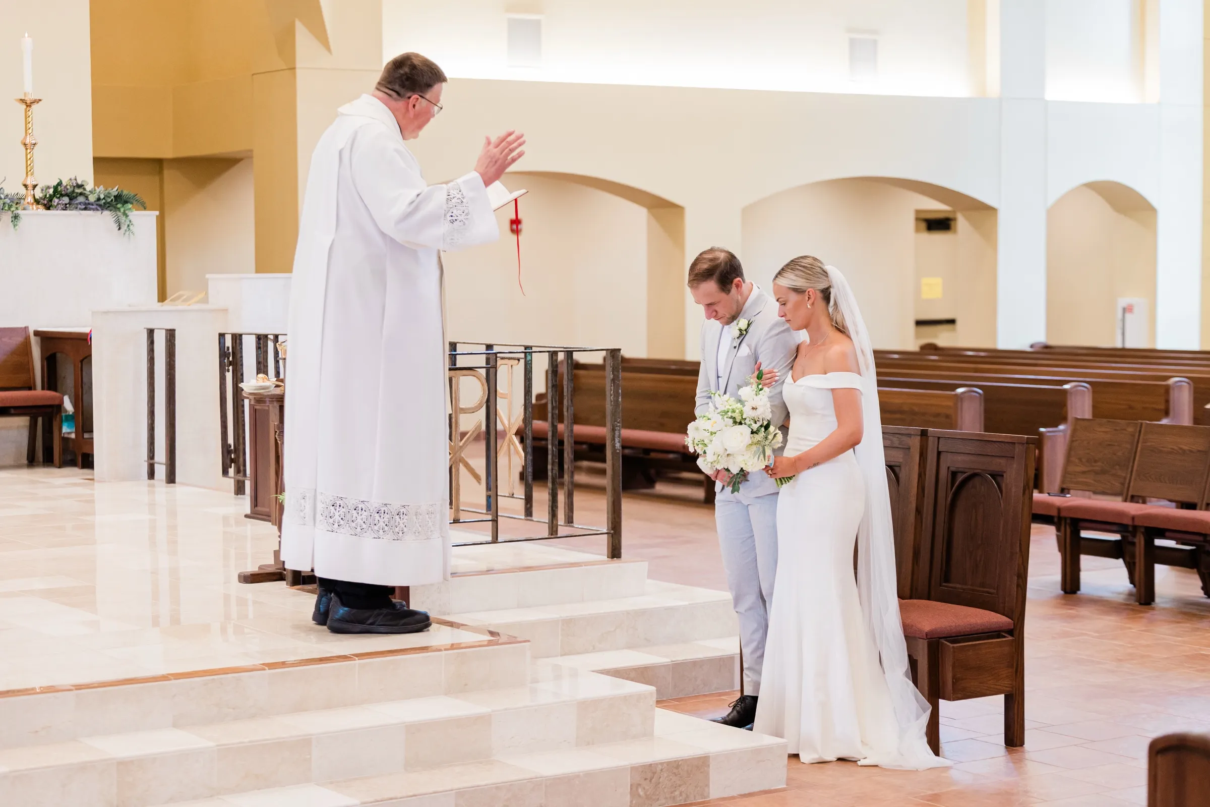 The priest raises his hand in blessing as Josse and Alex stand on the altar steps with heads bowed — Tim Larsen Photography, Brainerd Lakes MN