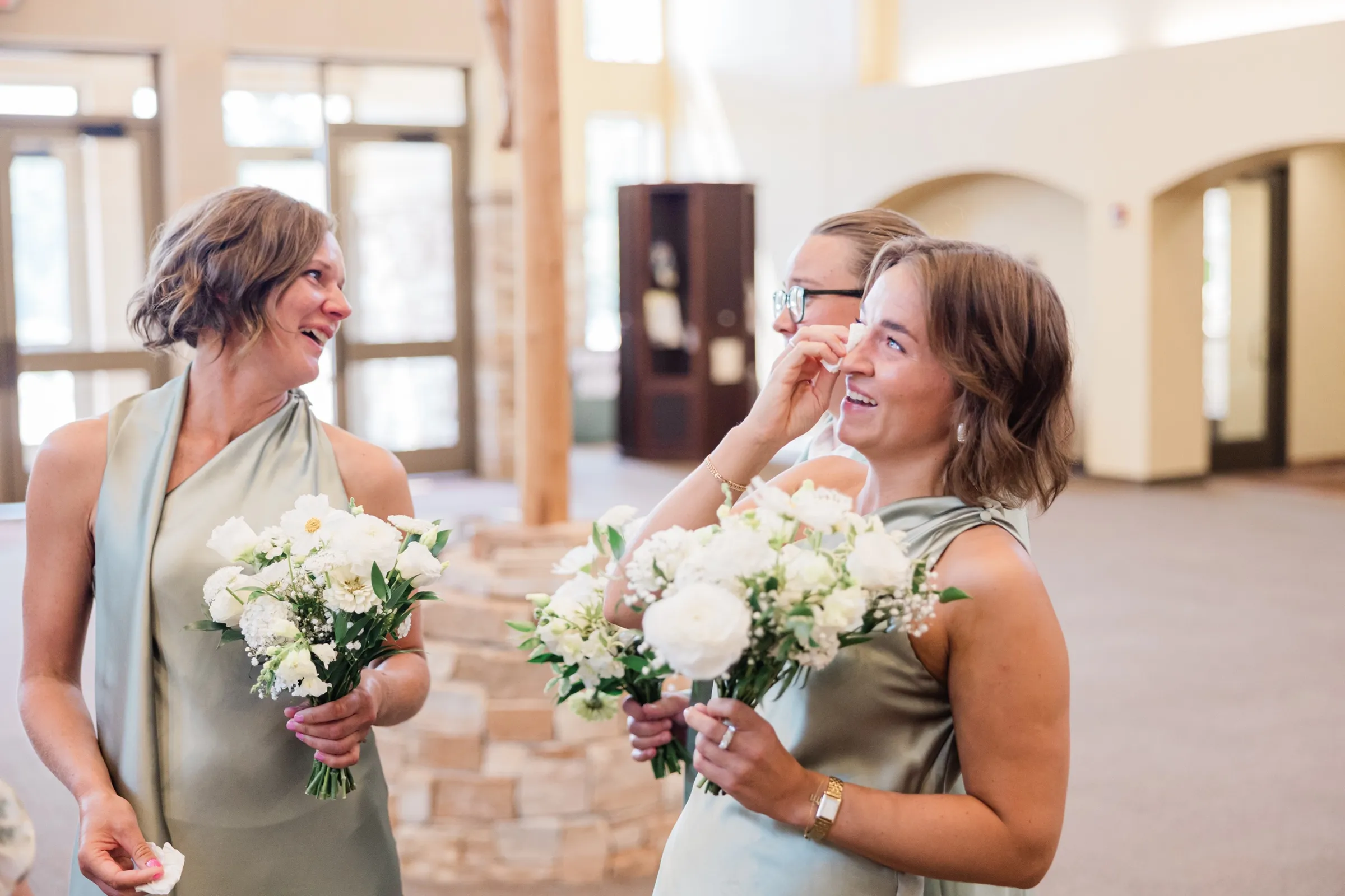 Three bridesmaids in sage green hold white bouquets in the church lobby, one wiping her eyes while another laughs — Tim Larsen Photography, Brainerd Lakes MN