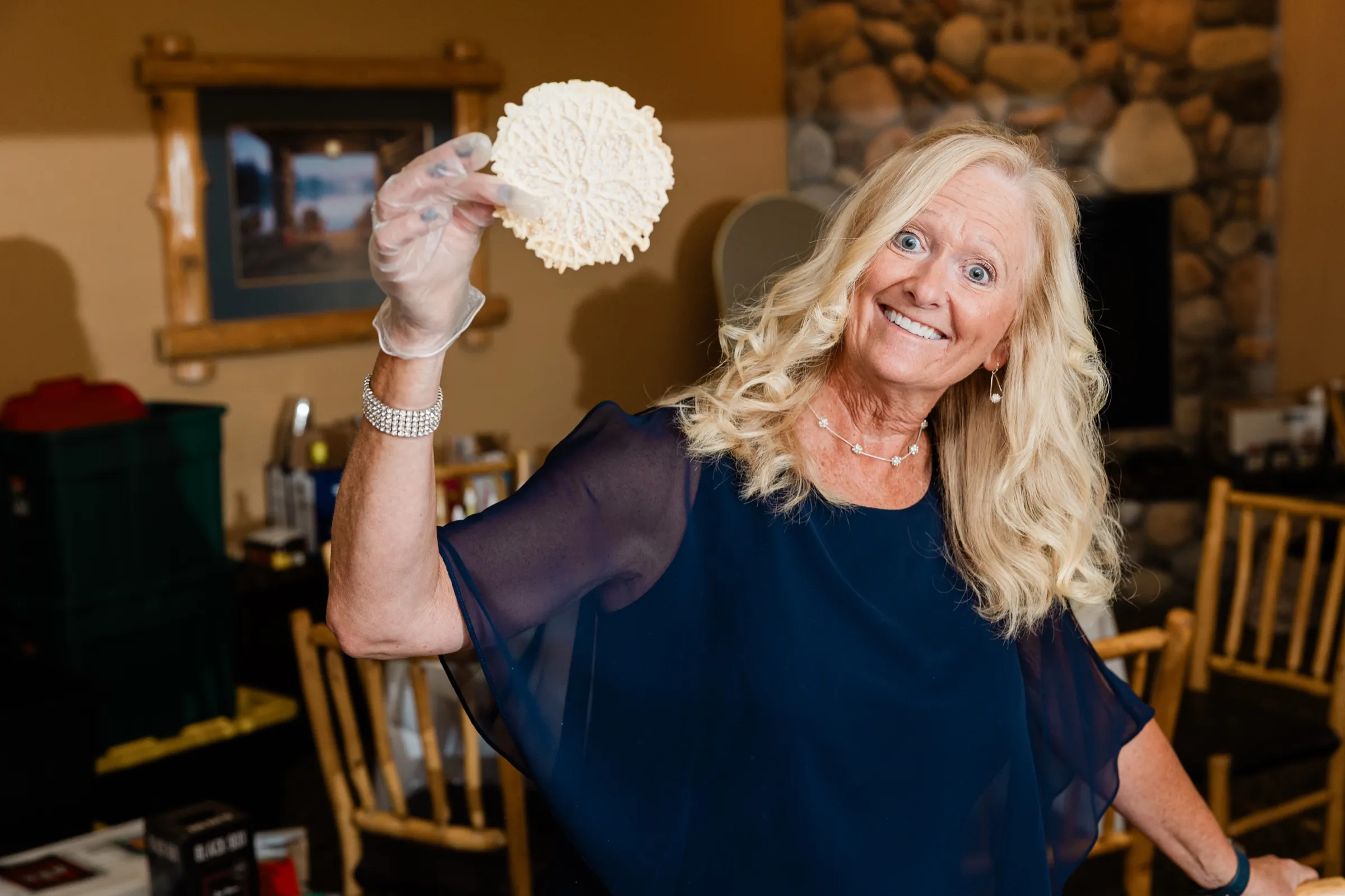 A guest in a navy dress holds up a lacy pizzelle cookie beside the fika table, a stone fireplace behind her — Tim Larsen Photography, Brainerd Lakes MN