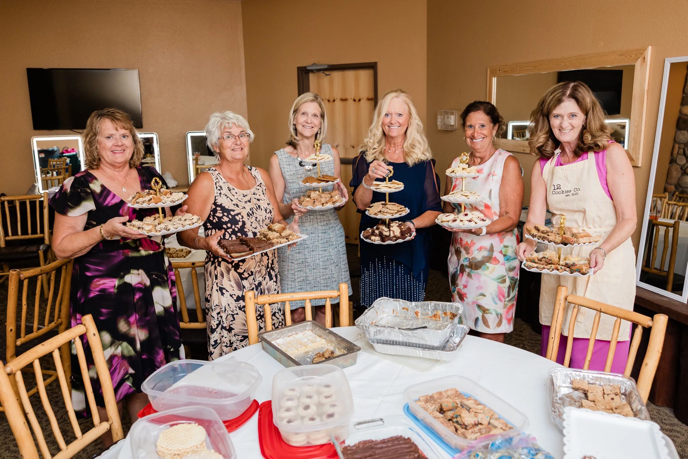 Six women stand shoulder to shoulder holding tiered plates of cookies and bars behind a table of homemade desserts, one in a baker's apron — Tim Larsen Photography, Brainerd Lakes MN