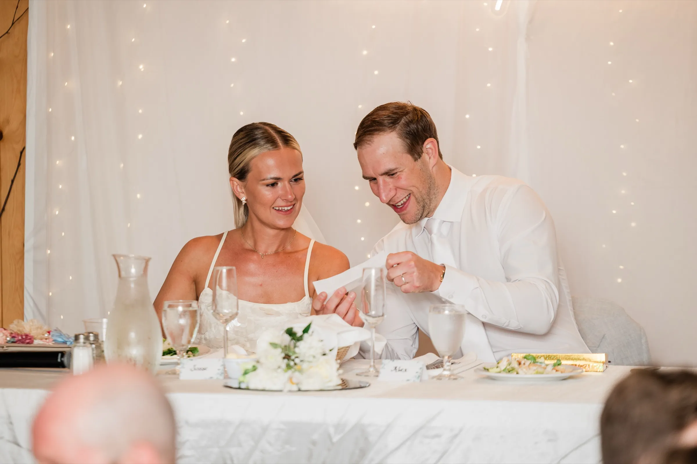 Seated at the head table, Alex laughs holding up a folded note as Josse reads along, string lights glowing behind them — Tim Larsen Photography, Brainerd Lakes MN