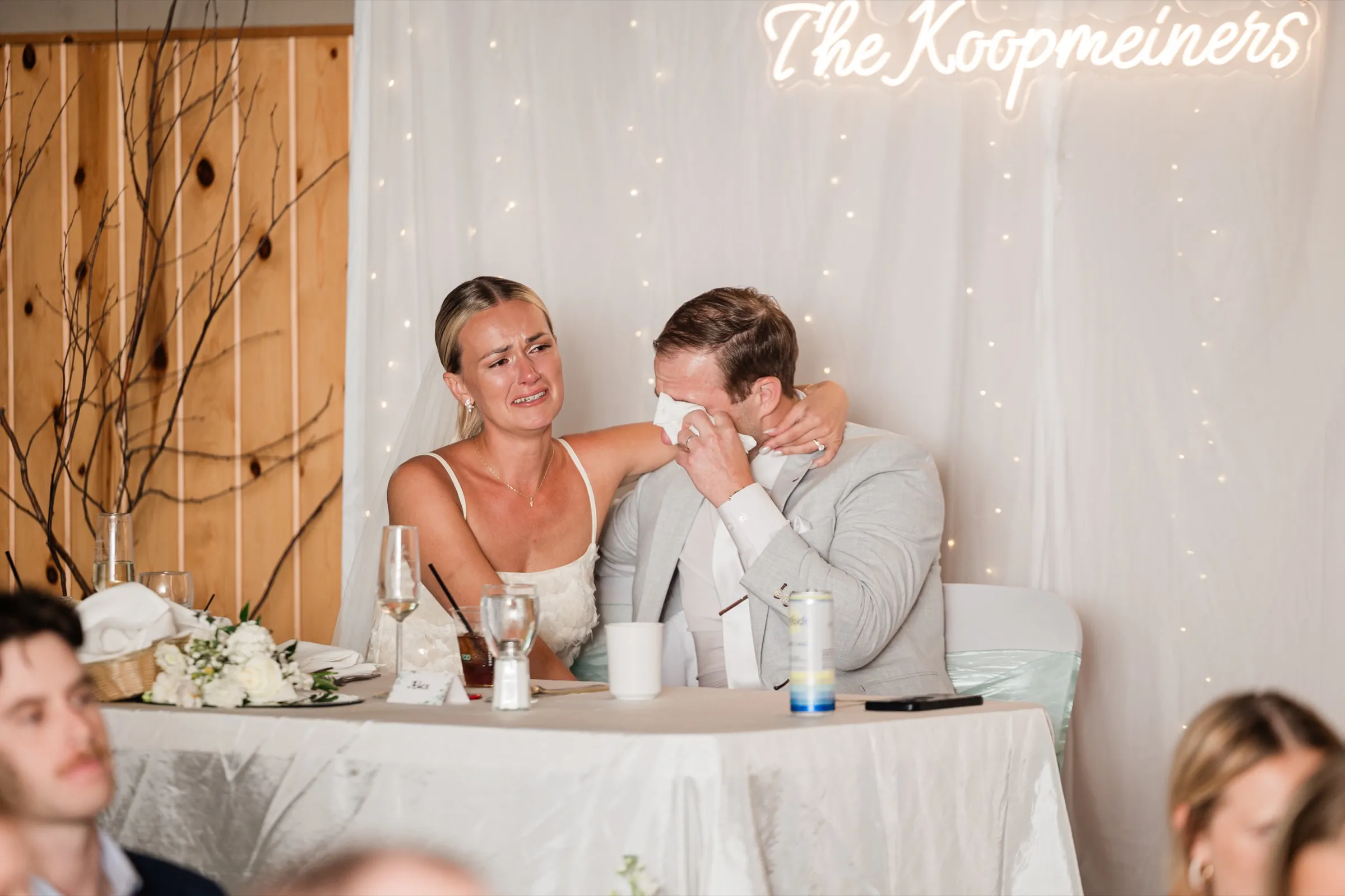 At the head table, Alex wipes his eyes with a napkin while Josse, also crying, holds him around the shoulders beneath the neon sign — Tim Larsen Photography, Brainerd Lakes MN