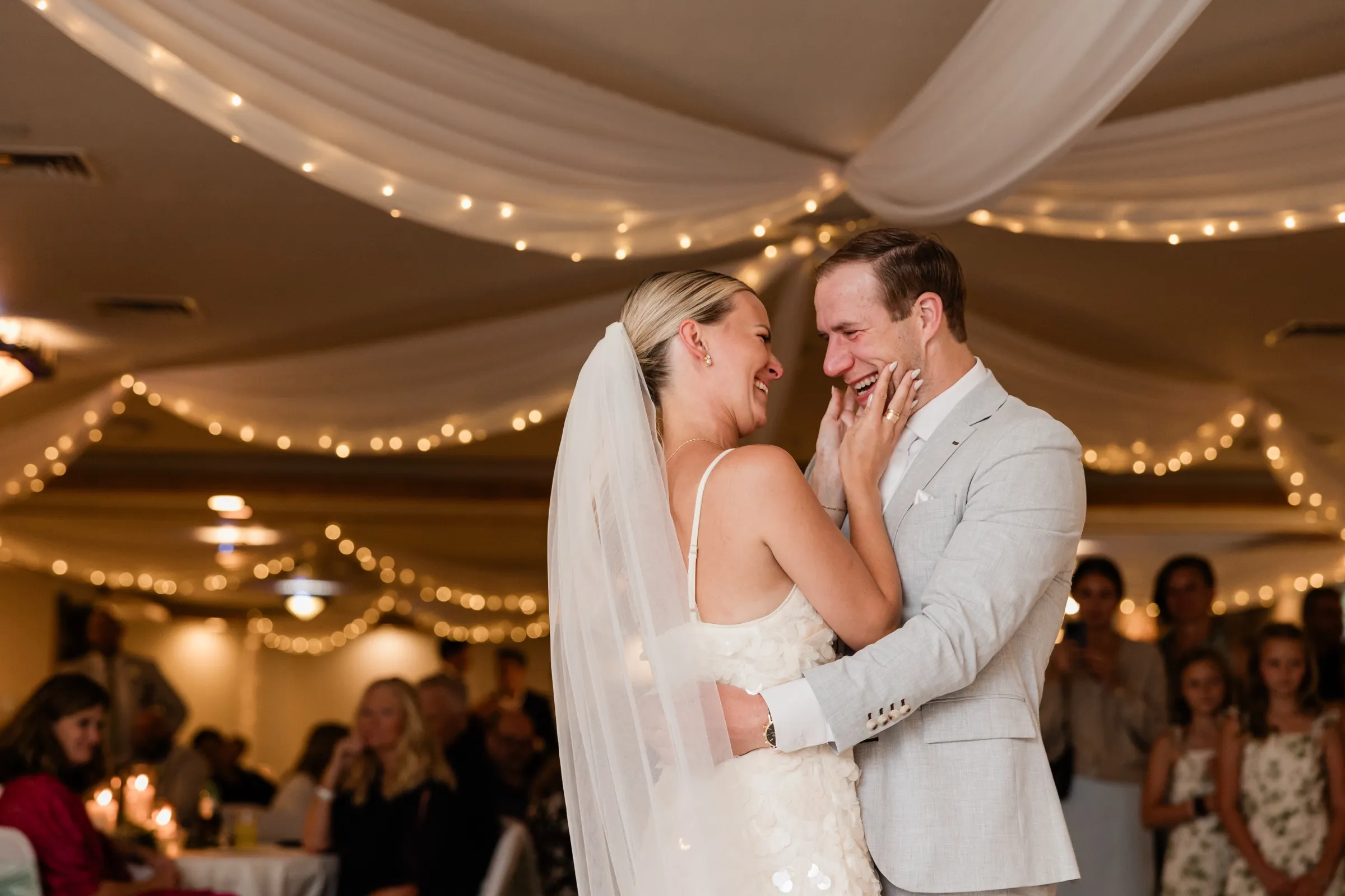 Josse cups Alex's cheek as they laugh face to face during their first dance beneath string lights at Whitefish Lodge in Crosslake — Tim Larsen Photography, Brainerd Lakes MN