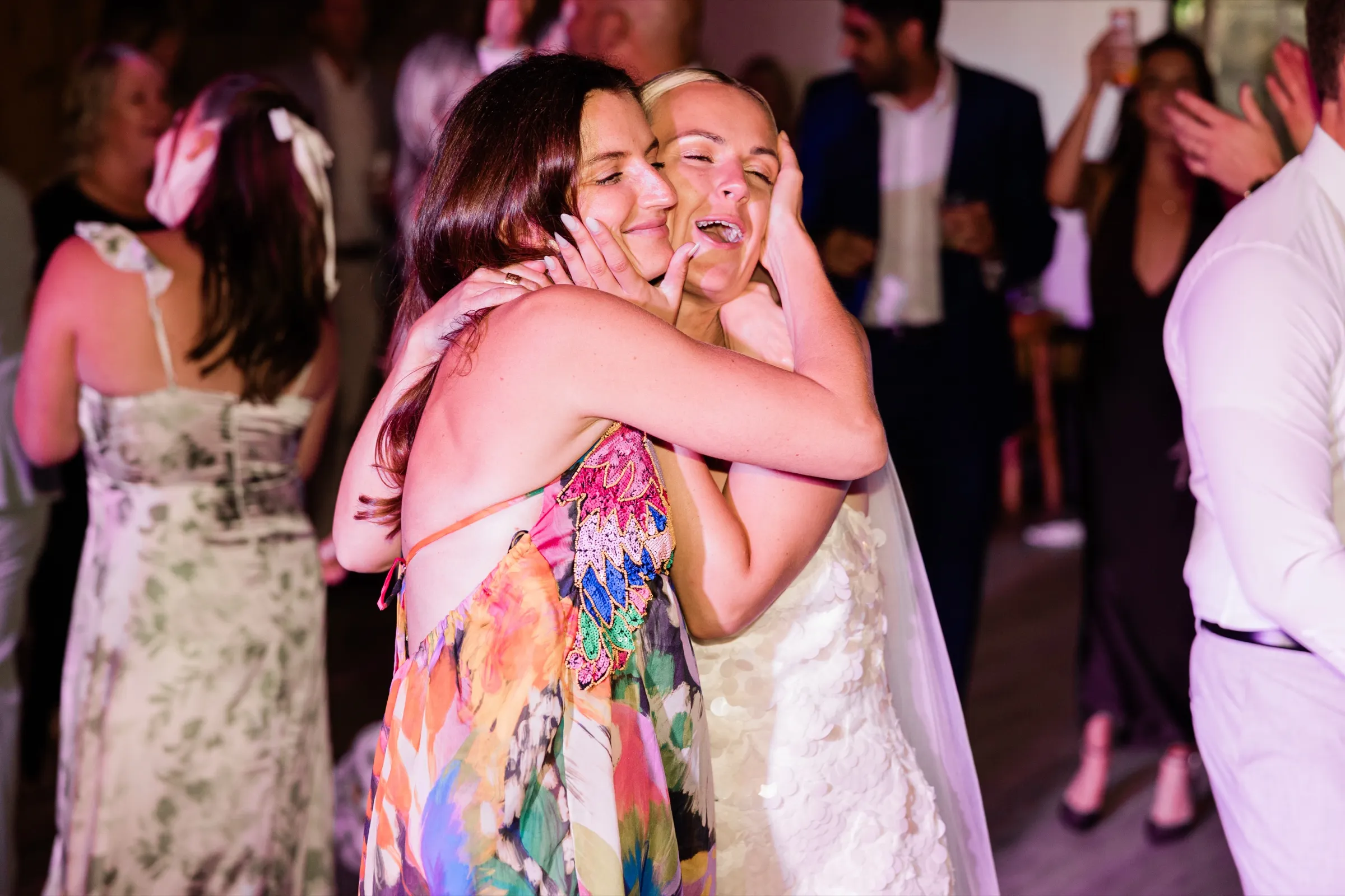 Josse laughs with her eyes closed as a guest in a floral dress holds her face and embraces her on a crowded dance floor — Tim Larsen Photography, Brainerd Lakes MN