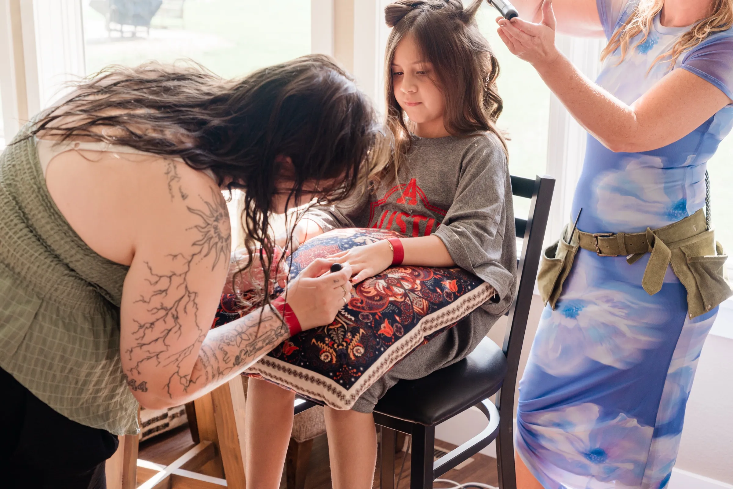 A young flower girl sits on a stool with a pillow in her lap while one woman paints her nails and another curls her hair. — Tim Larsen Photography, Brainerd Lakes MN