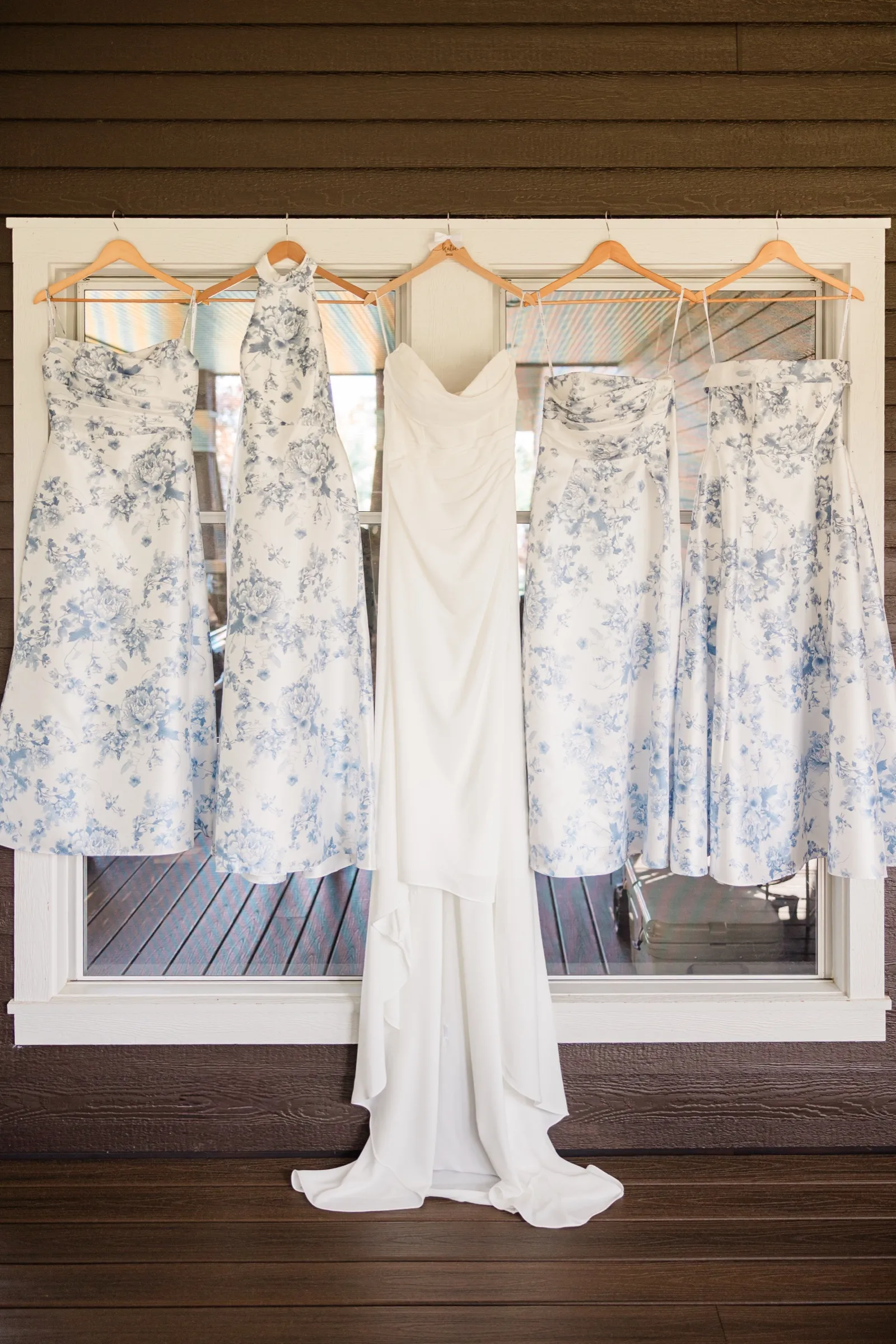 The bride's ivory gown hangs at the center of a cottage window flanked by four blue floral bridesmaid dresses. — Tim Larsen Photography, Brainerd Lakes MN