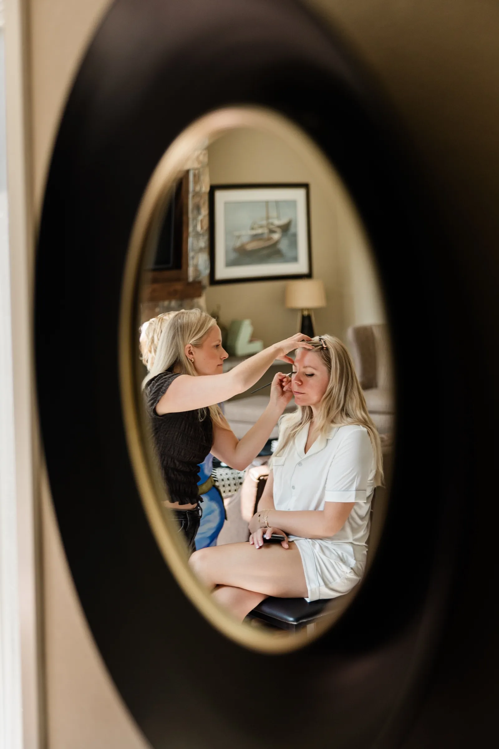 Reflected in a dark oval mirror, the bride sits with her eyes closed while the makeup artist finishes her face beneath a framed sailboat print. — Tim Larsen Photography, Brainerd Lakes MN