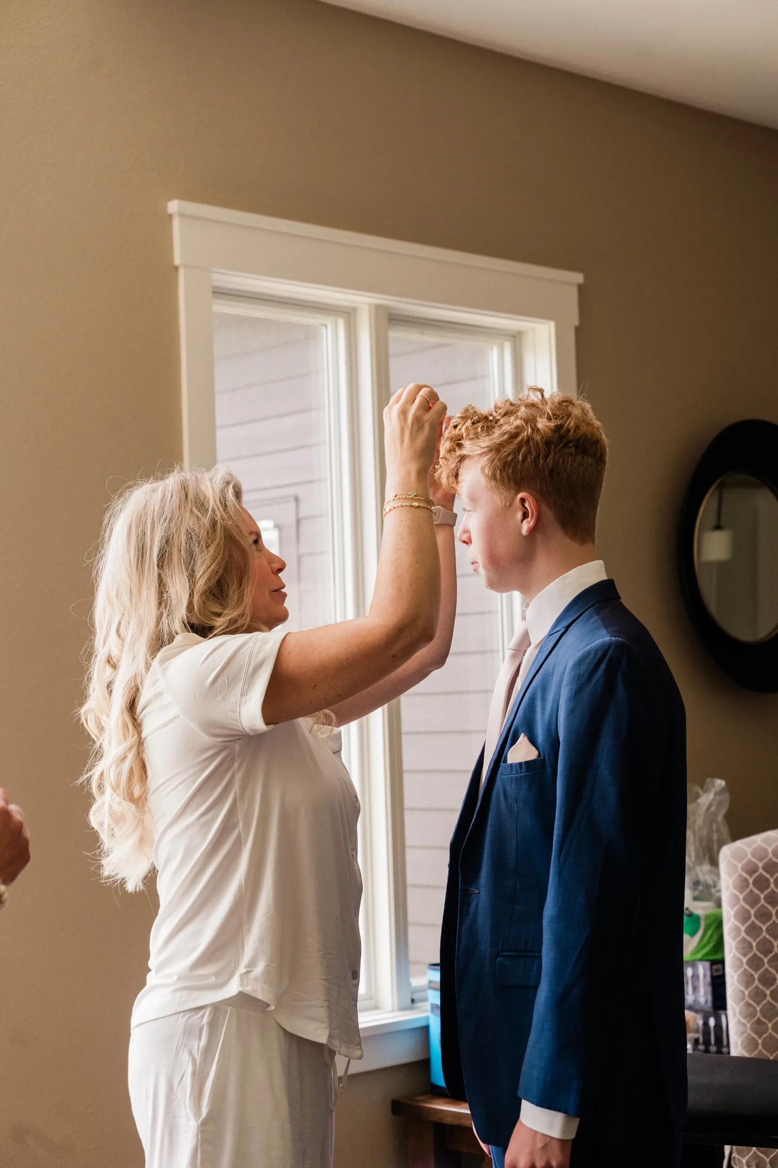 Still in her white pajamas, the bride reaches up to smooth her son's hair while he stands in his blue suit beside a window. — Tim Larsen Photography, Brainerd Lakes MN