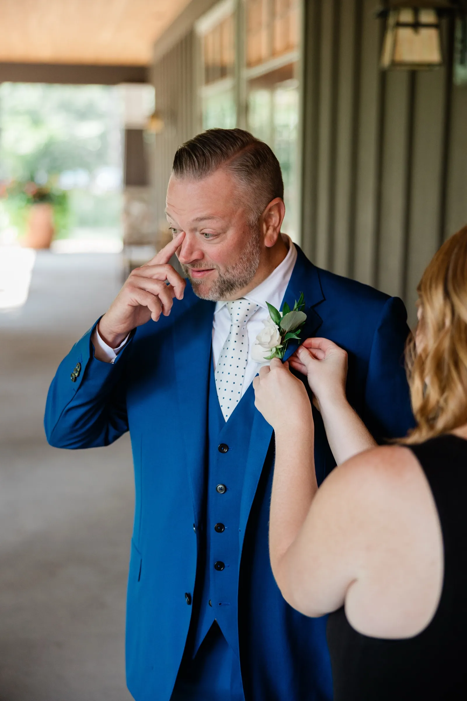 A woman in a black dress pins a white rose boutonniere to the groom's blue lapel on a shaded porch while he looks down at her hands. — Tim Larsen Photography, Brainerd Lakes MN