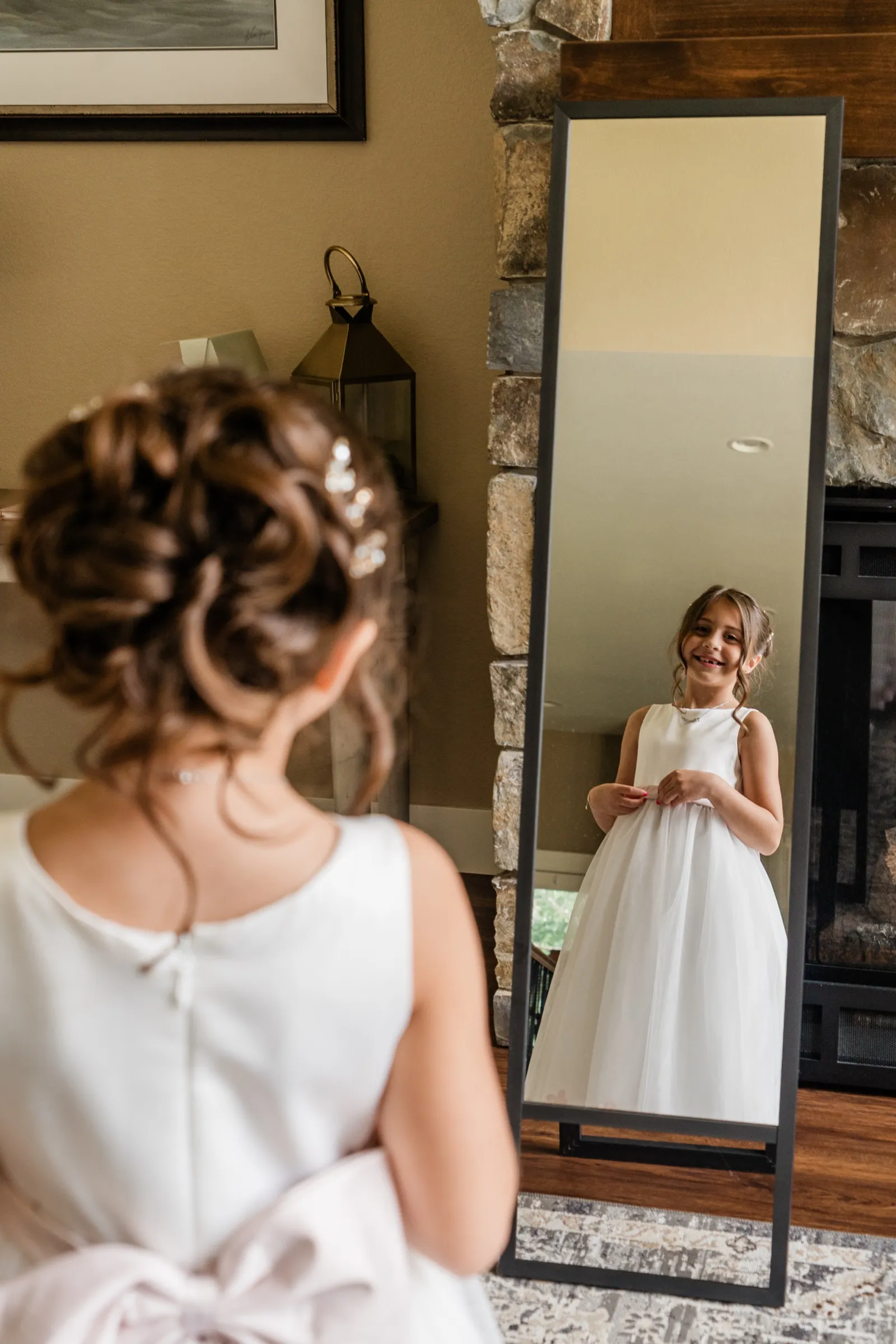 A flower girl in a white dress grins at herself in a standing mirror while an older girl with an updo watches from the blurred foreground. — Tim Larsen Photography, Brainerd Lakes MN