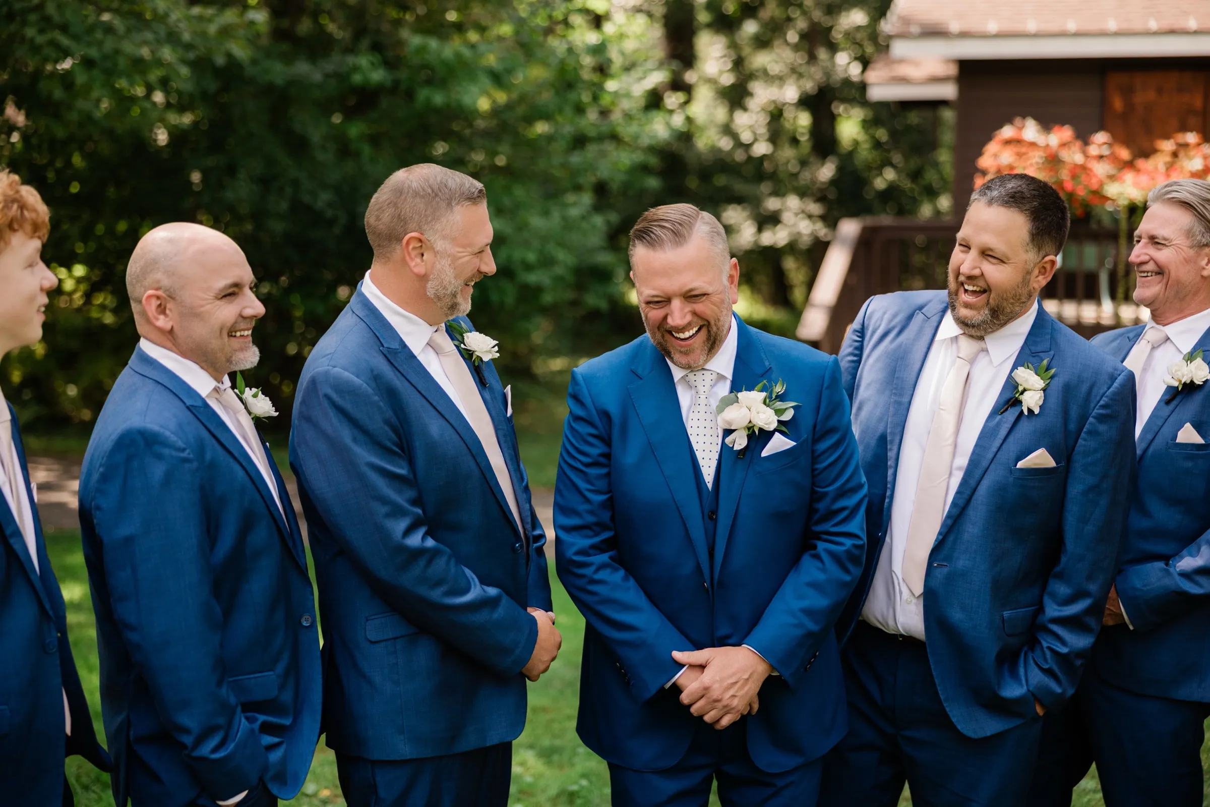 The groom laughs with his head lowered while six groomsmen in blue suits laugh with him on a shaded lawn. — Tim Larsen Photography, Brainerd Lakes MN
