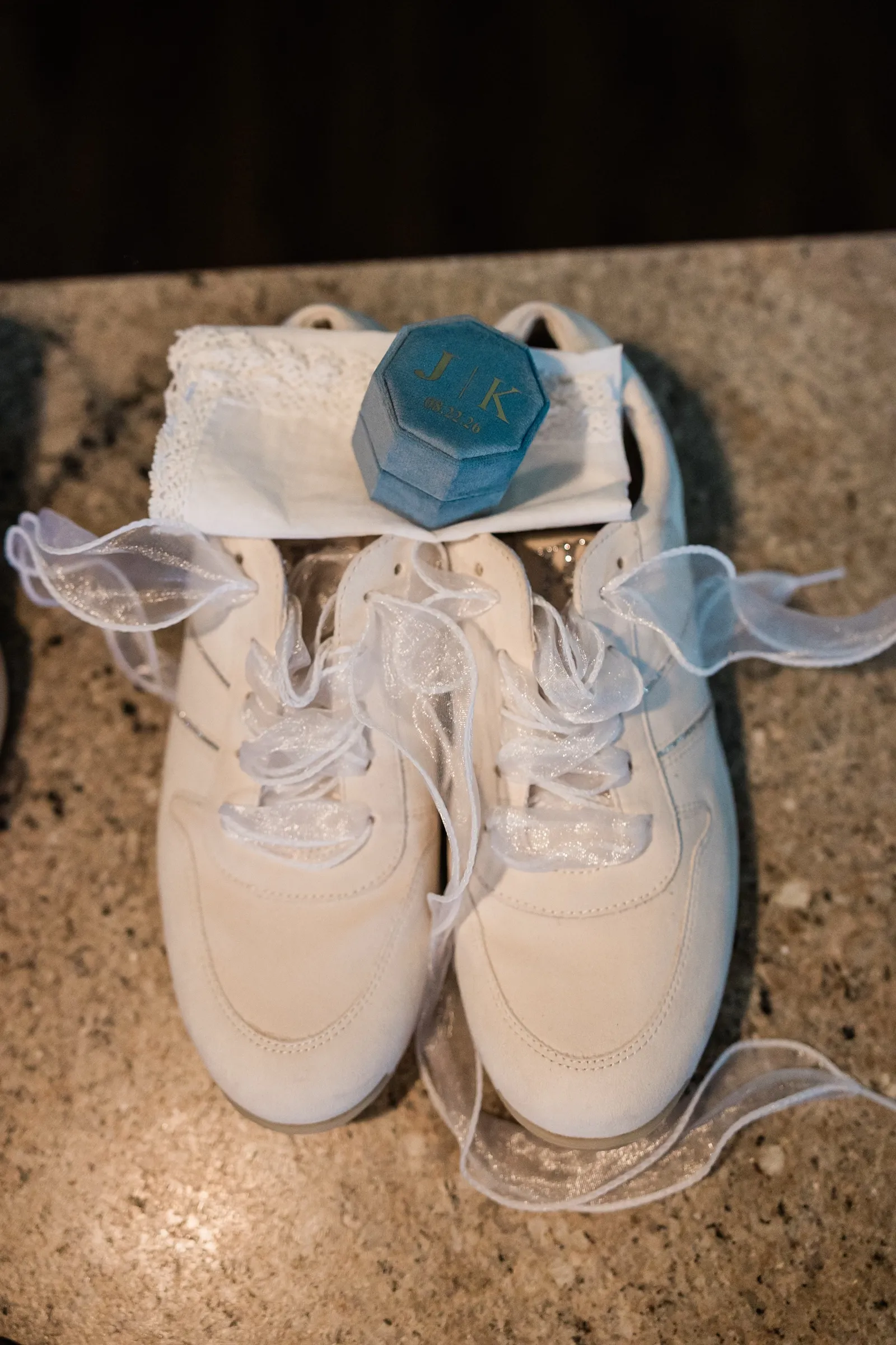 A pair of white bridal sneakers with sheer ribbon laces sits on a stone counter beside a blue velvet ring box monogrammed J and K with the wedding date. — Tim Larsen Photography, Brainerd Lakes MN