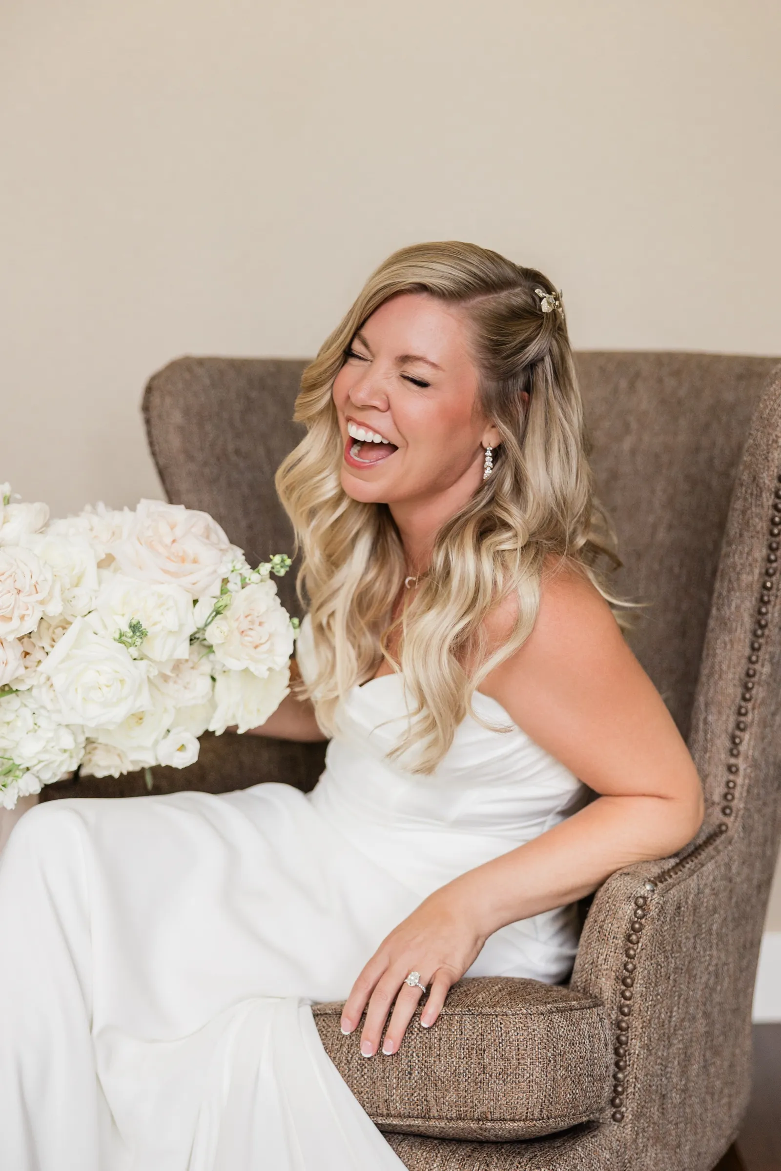 The bride laughs with her head tipped back and her eyes closed while seated in a wingback chair, her bouquet held out to one side. — Tim Larsen Photography, Brainerd Lakes MN