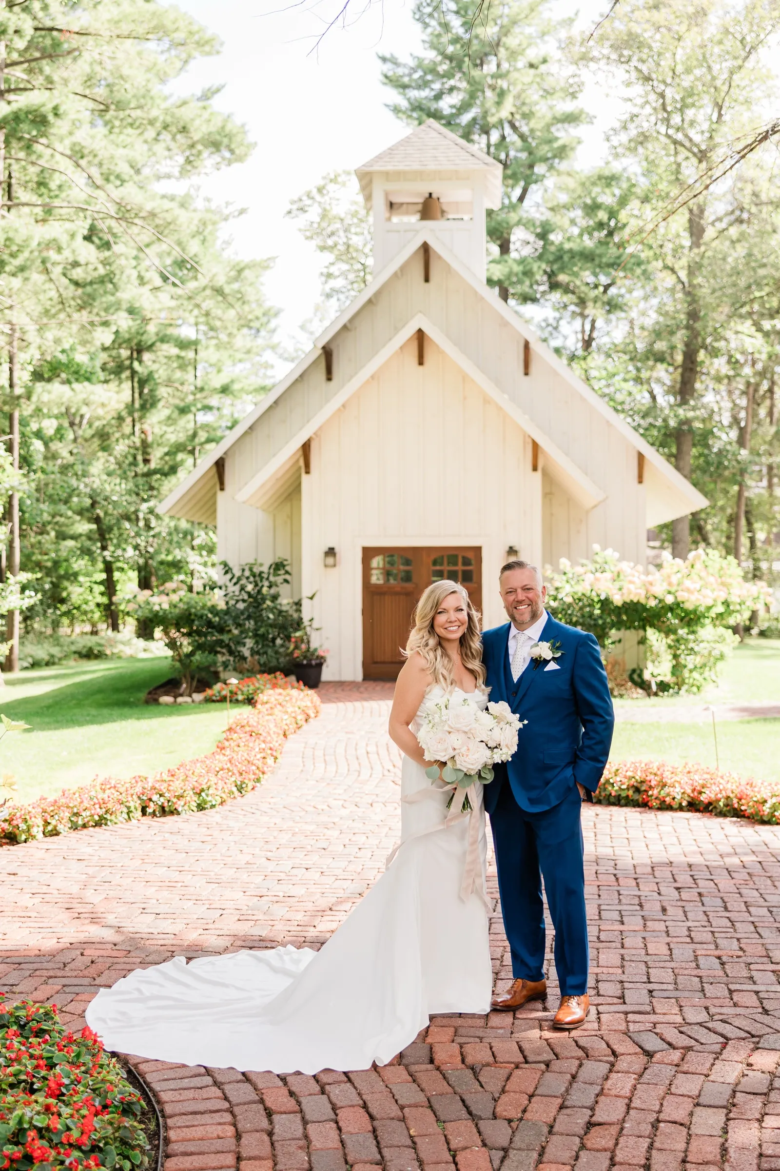 The bride and groom stand together on a curved brick courtyard in front of a white wooden chapel with a bell tower, flower beds lining the path. — Tim Larsen Photography, Brainerd Lakes MN